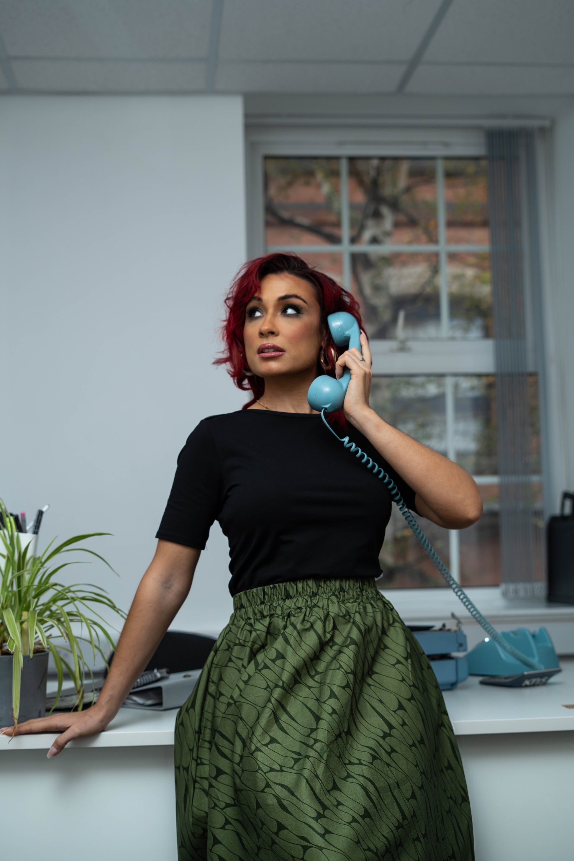 Woman in a black top and green skirt talking on a blue phone in an office setting.