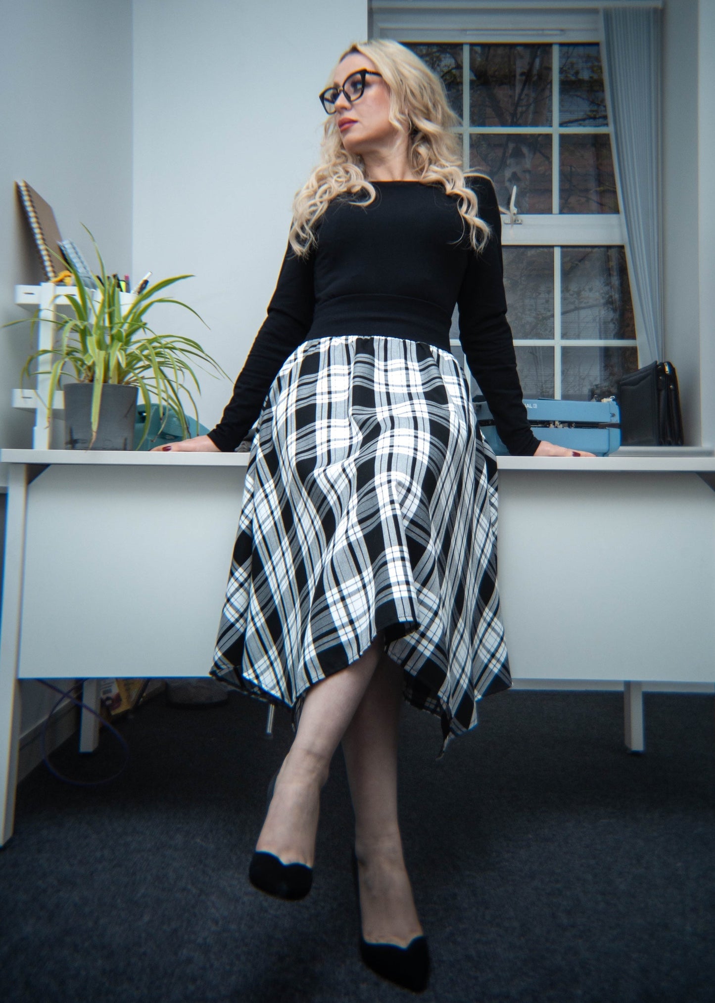Woman sitting at a desk in an office wearing a black top and plaid skirt.