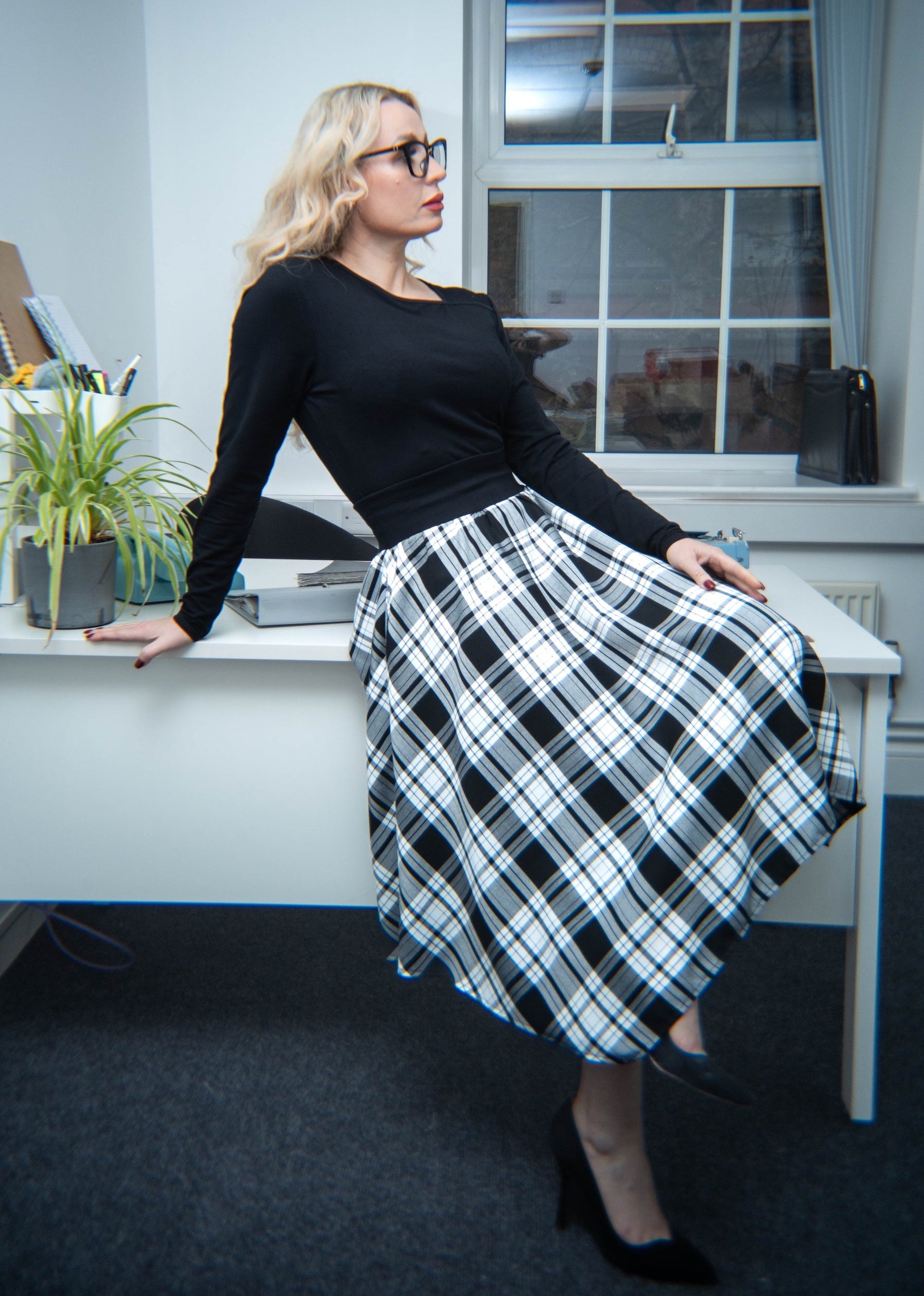 Woman sitting at a desk in an office setting wearing a black top and plaid skirt.