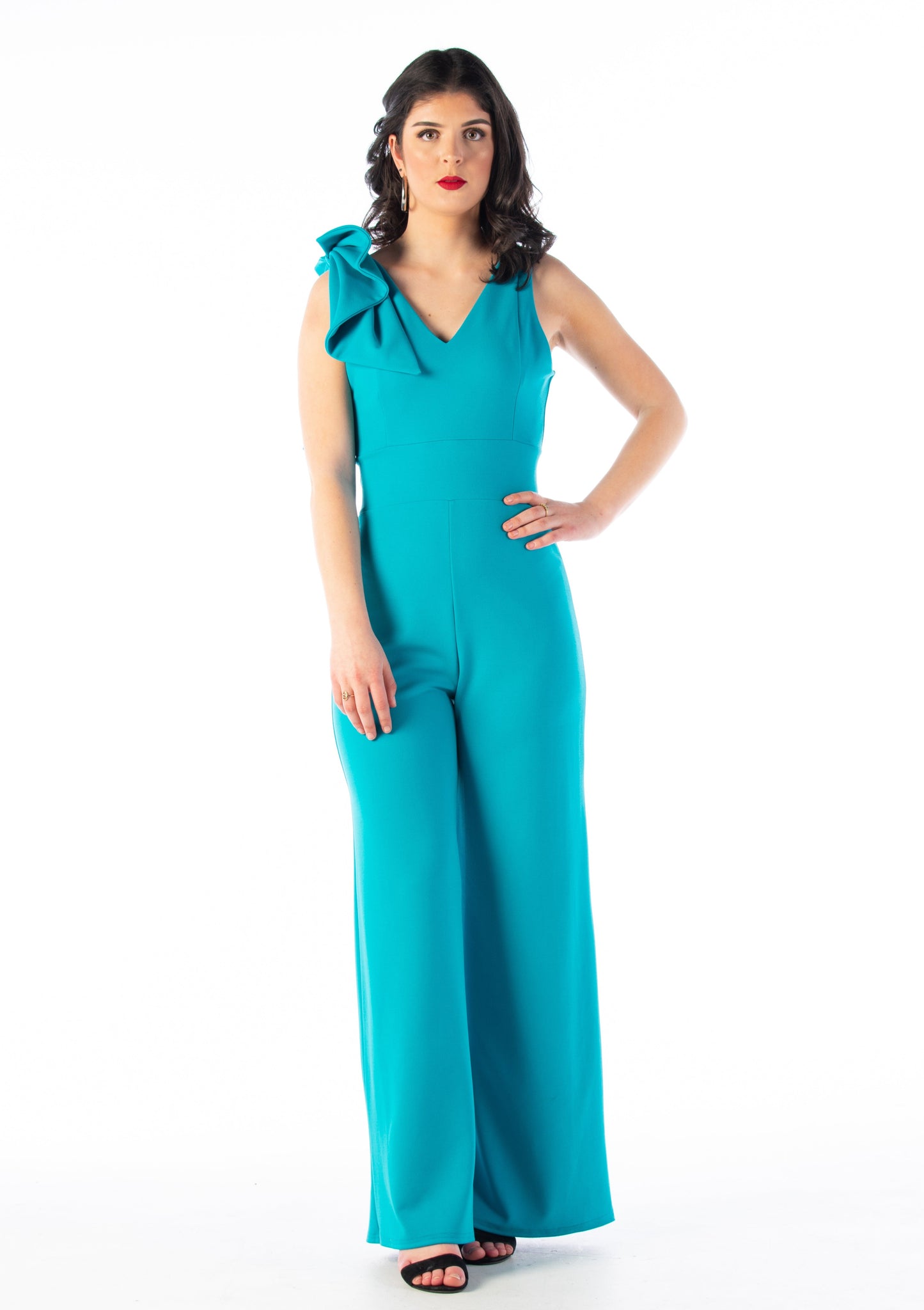 This is a young lady wearing a Turquoise colour V neck jumpsuit with wide legs and a bow on the shoulder. She is wearing black strappy heels and is standing with her hand on her hip, silhouetted against a white wall.