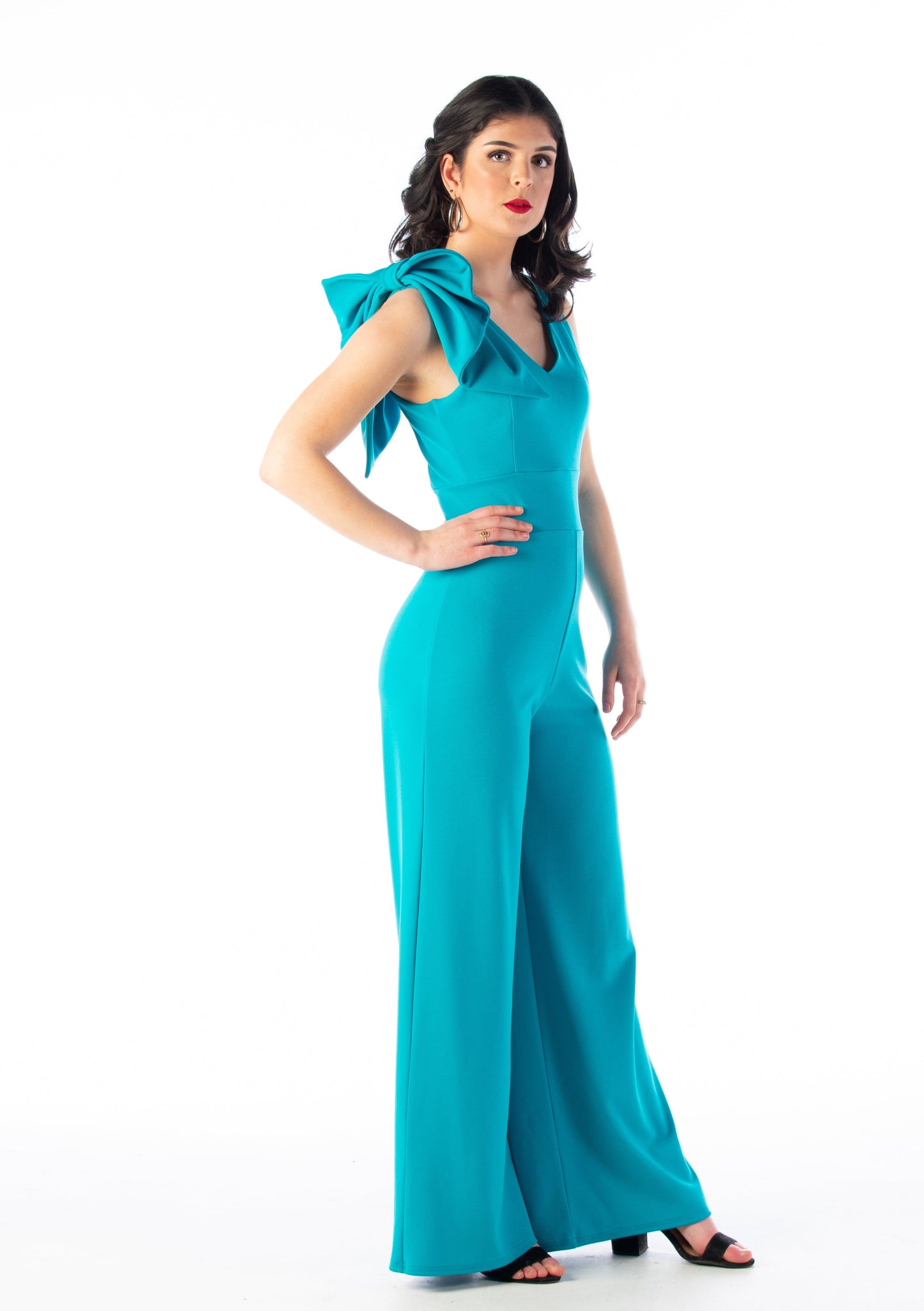 This is a young lady wearing a Turquoise colour V neck jumpsuit with wide legs and a bow on the shoulder. She is wearing black strappy heels and is standing with her hand on her hip, silhouetted against a white wall.