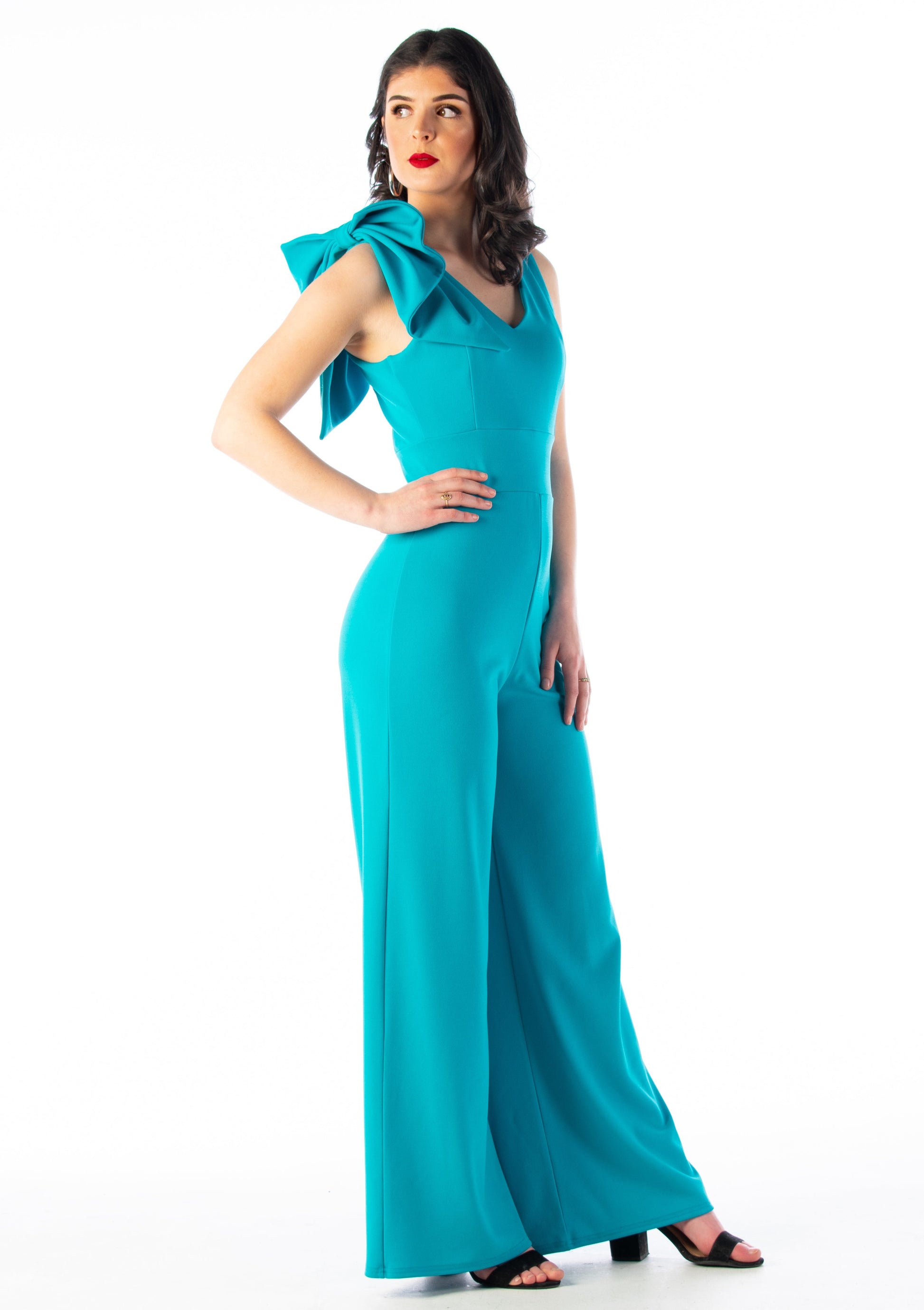 This is a young lady wearing a Turquoise colour V neck jumpsuit with wide legs and a bow on the shoulder. She is wearing black strappy heels and is standing with her hand on her hip and looking over her shoulder, silhouetted against a white wall.