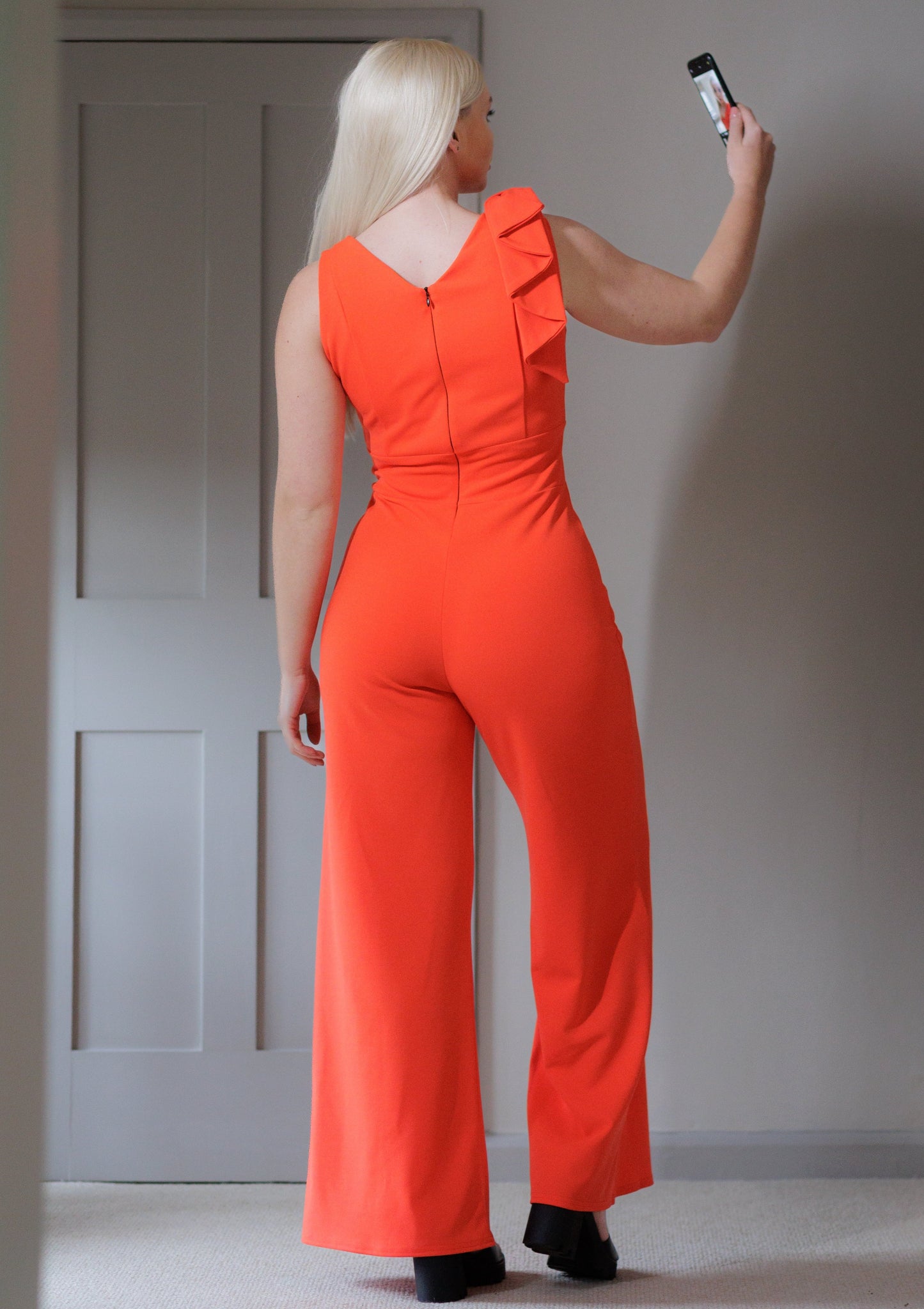 This is the back view a young lady wearing a Flame red coloured V neck jumpsuit, with wide legs and a bow on one shoulder. She is wearing black open-toed sandals and is taking a selfie on a black phone.