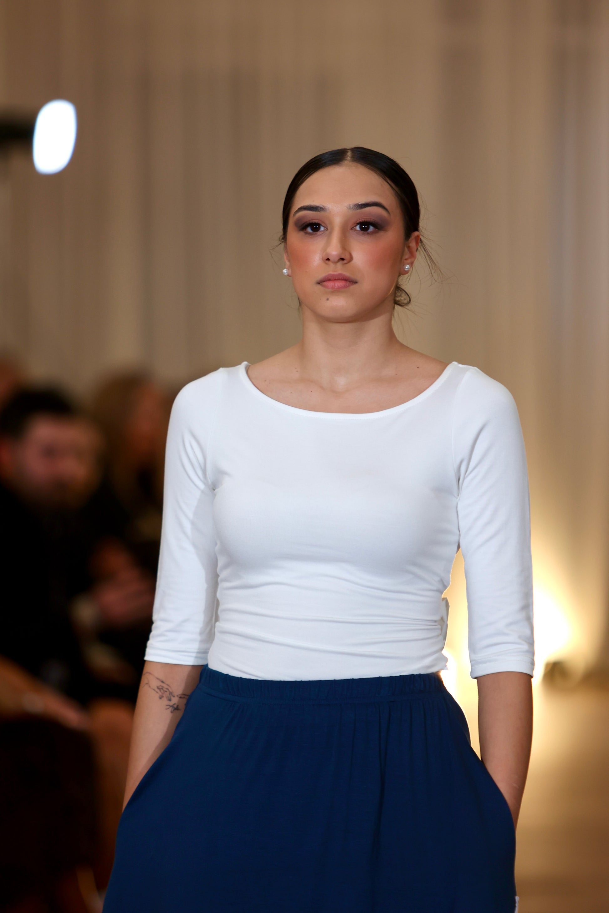 This is a young woman facing the camera silhouetted against a cream drape background. Her hair is tied back, she wears a cream 3/4 length sleeve top with a scoop neck. She wears a our blue Boho Skirt with is which has pockets and a deep gathered frill.