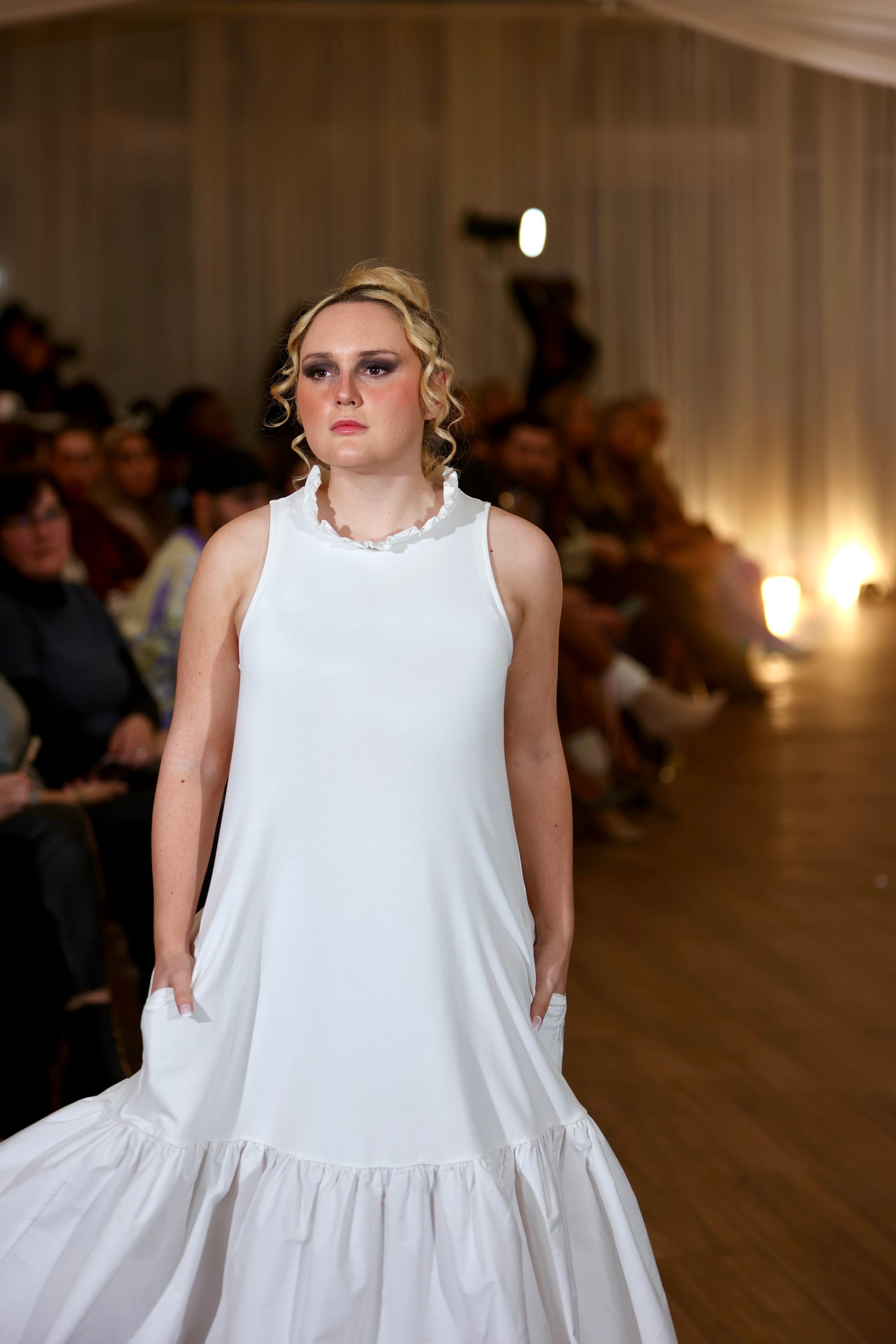 There is a young woman walking towards the camera in a dimly lit room  with people seated in the background. She has long blonde hair tied up. She is wearing the iris midi dress with pockets in cream. The dress has a high neck with a row of ruffles. Both hands are in her pockets. 