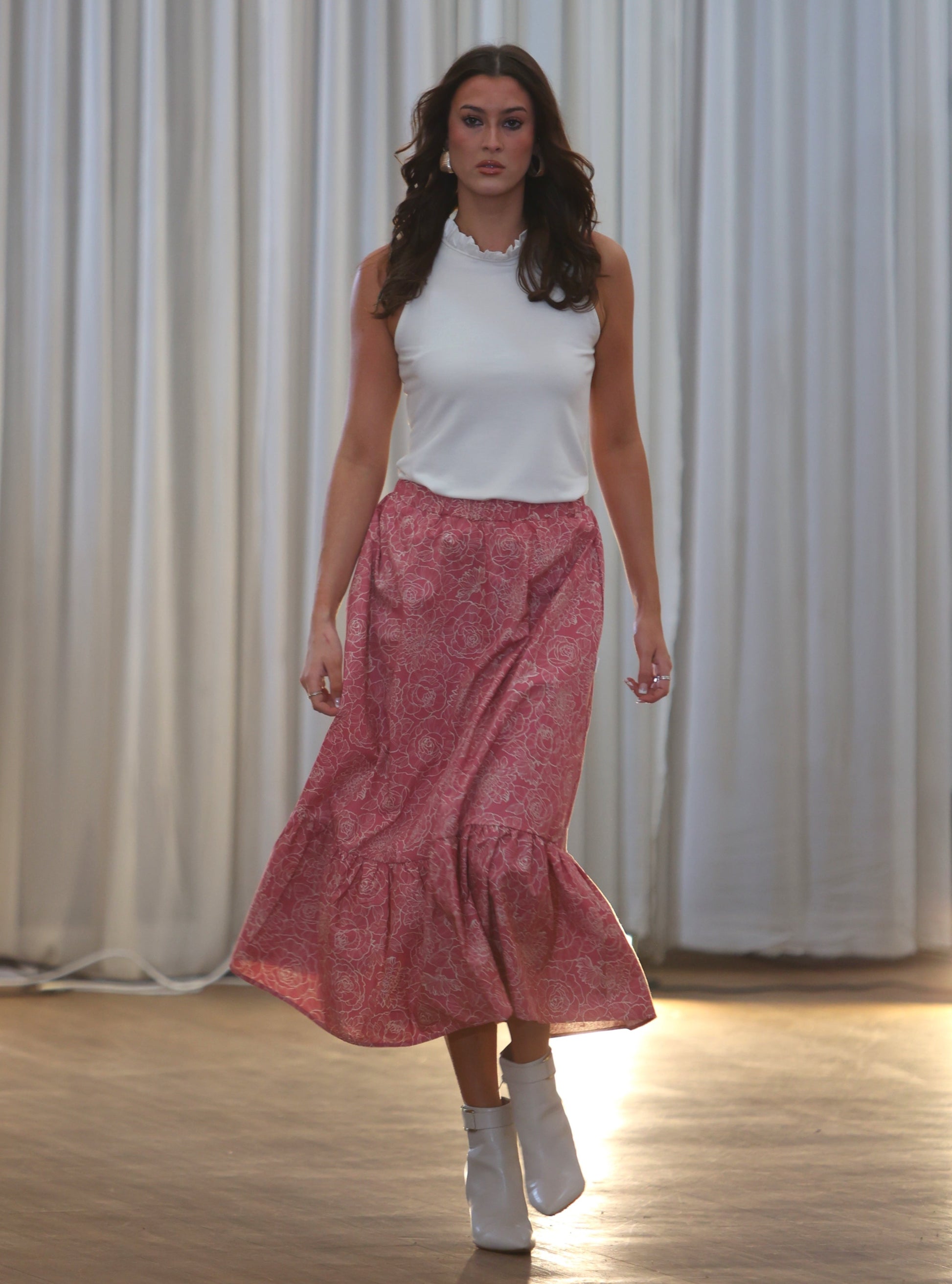This is a young woman walking towards the camera. She is wearing a pink and cream patterned skirt silhouetted against a white drape background. It has an elasticated waist and frill hem. She wears this with a cream sleeveless top with ruffle neck.
