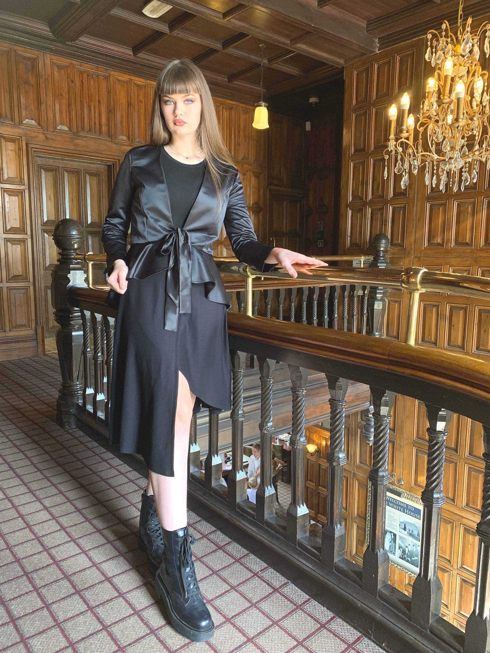 Our model faces the front but is looking aforward at the camera. She is holding the banister with her left hand and the hem of the jacket in her right hand. Her long dark hair is falling behind her down her back. She is on a landing with dark wood  panelling and a brass banister. She is wearing a mock wrap skirt that has an asymmetrical hemline with a black crew neck long sleeved bodysuit.