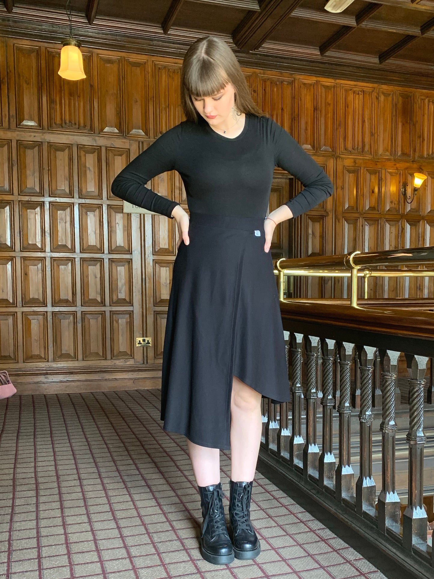 Our model faces the front but is looking down to the floor. She has her hands on her hips and her long dark hair is falling behind her down her back. She is on a landing with dark wood  panelling and a brass banister. She is wearing a mock wrap skirt that has an asymmetrical hemline with a black crew neck long sleeved bodysuit.