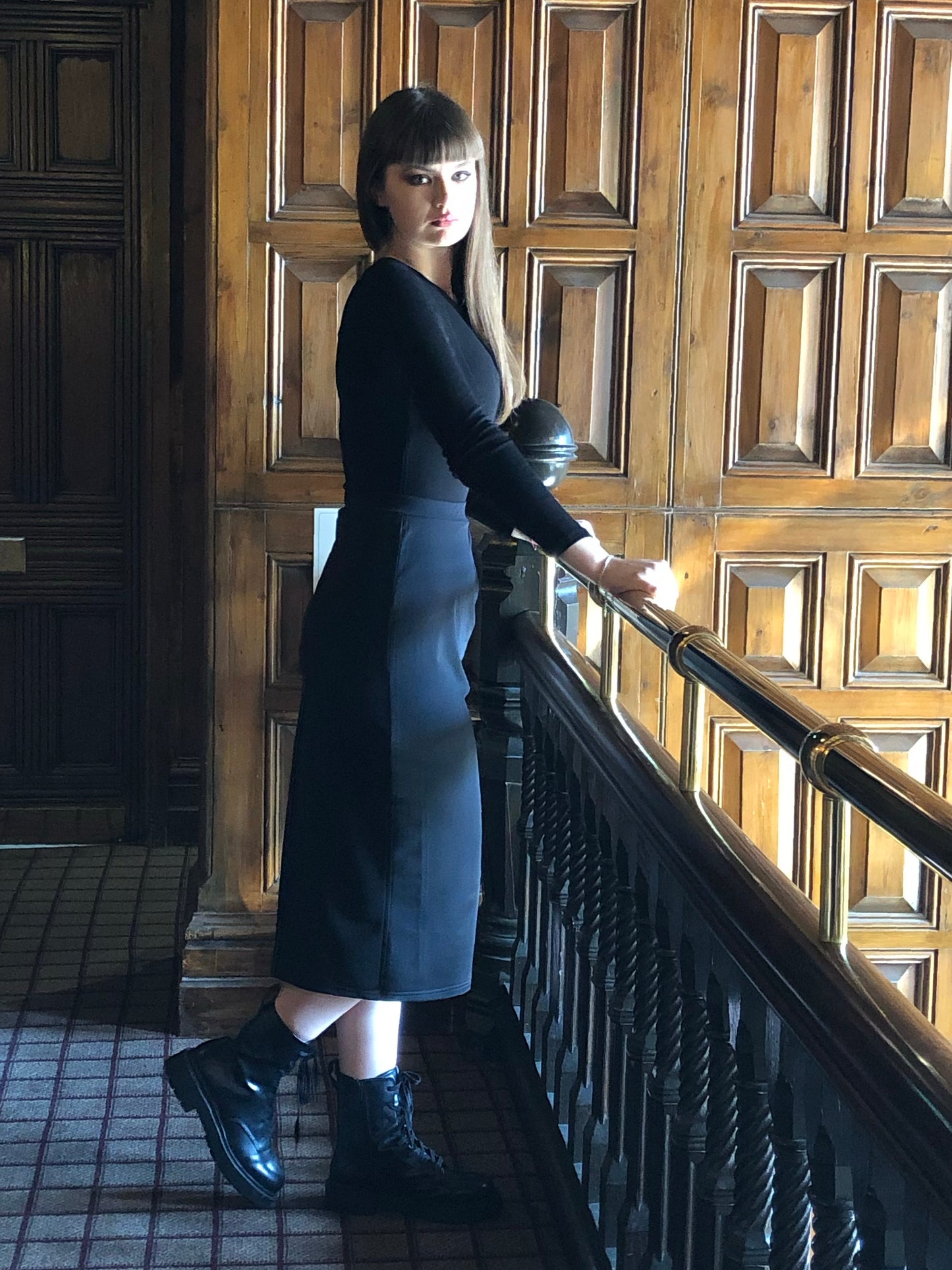 This is a young woman wearing a black long sleeve t-shirt with a below the knee, pencil style skirt, with a pleated waist. She has long brown hair and is wearing shiny black boots. She is silhouetted against a dark wood panelled room, leant against a banister, but looking over her shoulder, towards us.