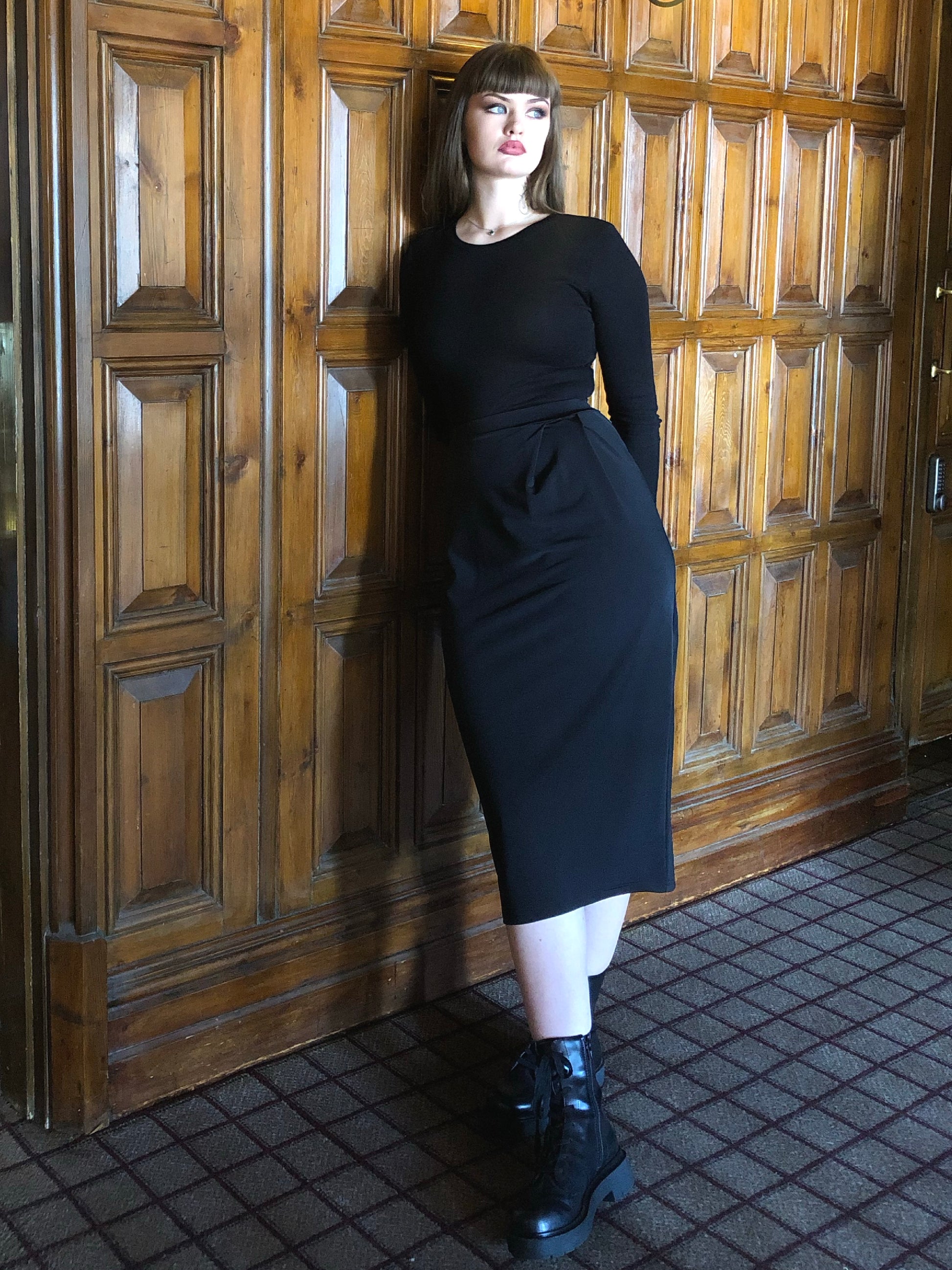 This is a young woman wearing a black long sleeve t-shirt with a below the knee, pencil style skirt, with a pleated waist. She has long brown hair and is wearing shiny black boots. She is silhouetted against a dark wood panelled room.