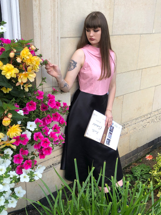 This is an A Line dress sleeveless with a boat collar. The top of the dress is baby pink with sparkle and the skirt is a structured dull duchess satin in black. It is worn by a model with long dark hair who is holding  a book, touching a flower and leaning against a wall. There are flowers to the left of the picture and also the edge of a window in the wall.