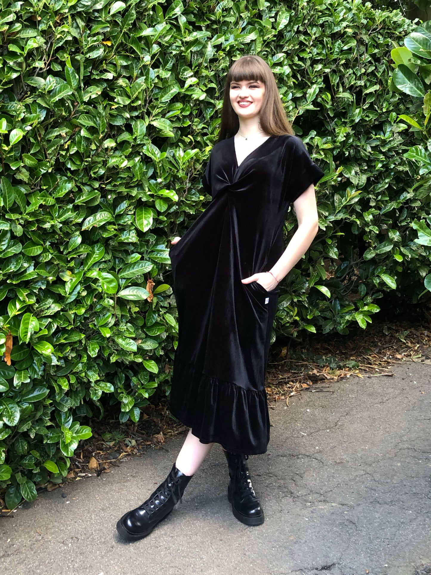 This is a black frill velvet dress worn by a model with dark hair against a Japonica hedge. It has a knot and plunge v neck with short sleeves. The model is facing front and has her hands in her pockets. She is wearing black boots.