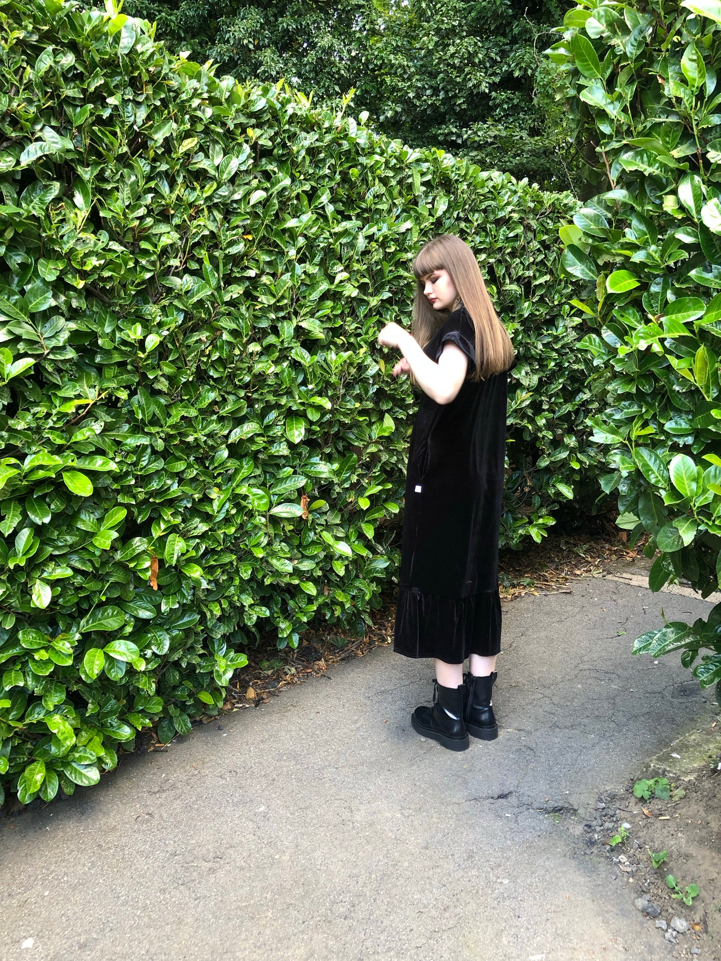 This is a black frill velvet dress worn by a model with dark hair against a Japonica hedge. It has a knot and plunge v neck with short sleeves. The model is facing the left side as you view the image. She is looking at a leaf and feeling it with her left hand. She is wearing black boots.