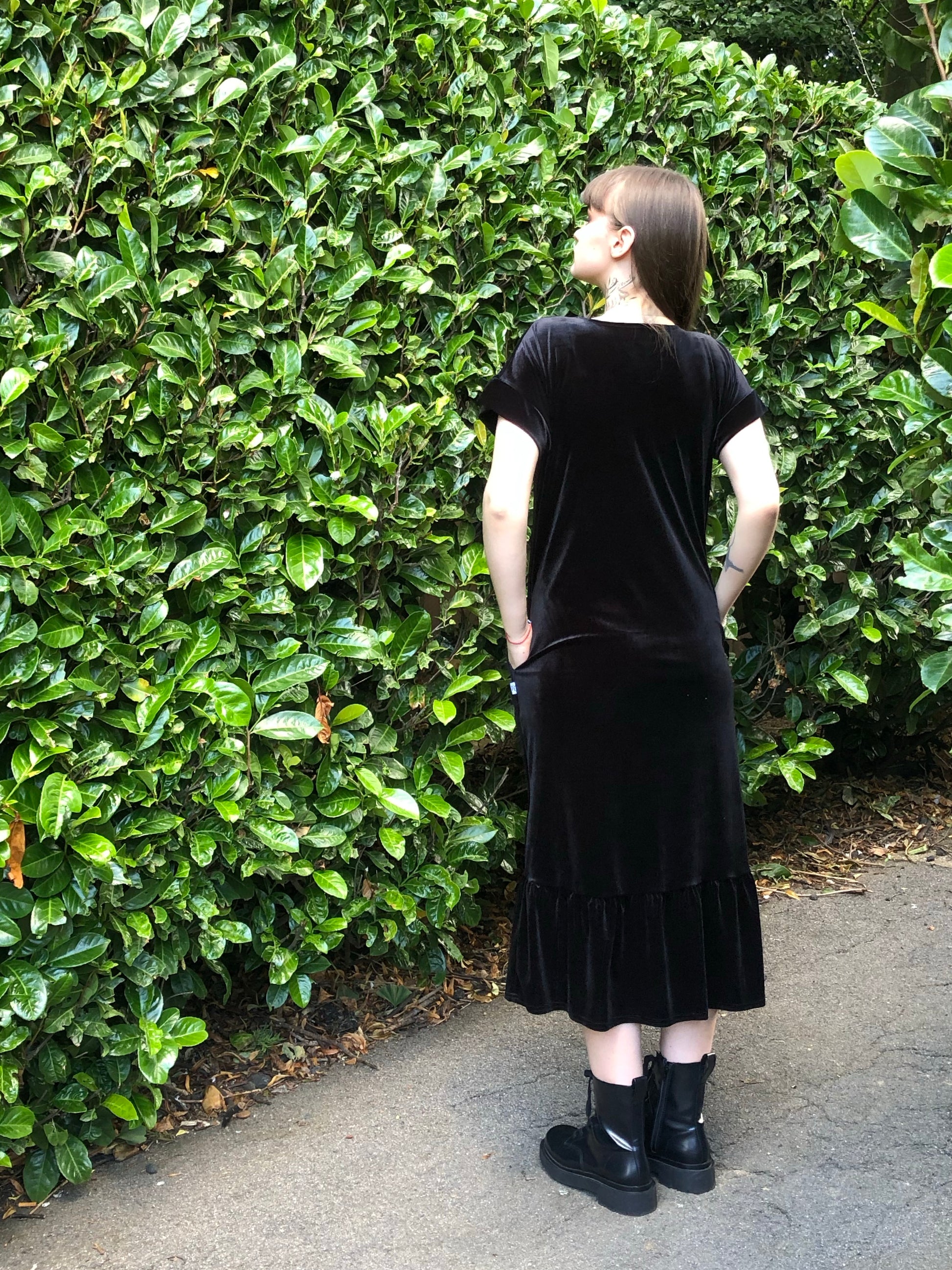 This is a black frill velvet dress worn by a model with dark hair against a Japonica hedge. It has a knot and plunge v neck with short sleeves. The model is facing the hedge and this is a back view of the dress and has her hands in her pockets. She is wearing black boots.