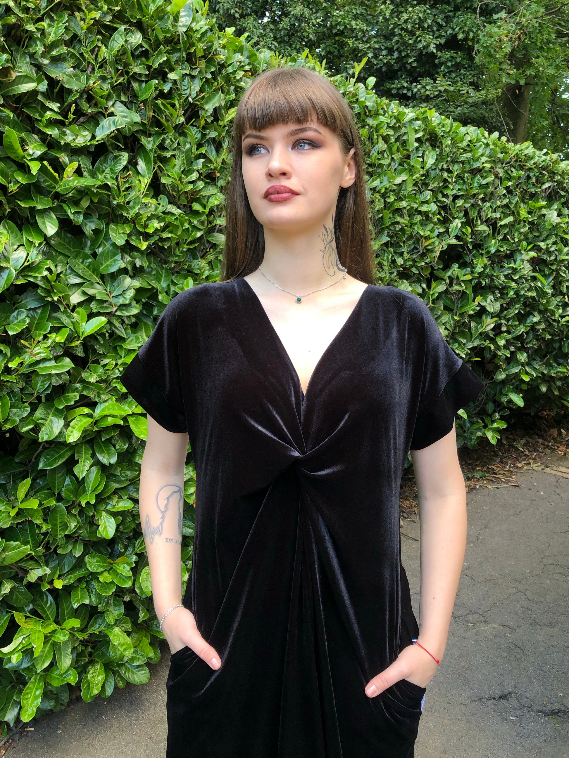 This is a black frill velvet dress worn by a model with dark hair against a Japonica hedge. The image is cut off at the top of her legs. It has a knot and plunge v neck with short sleeves. The model is facing front and has her hands in her pockets. 