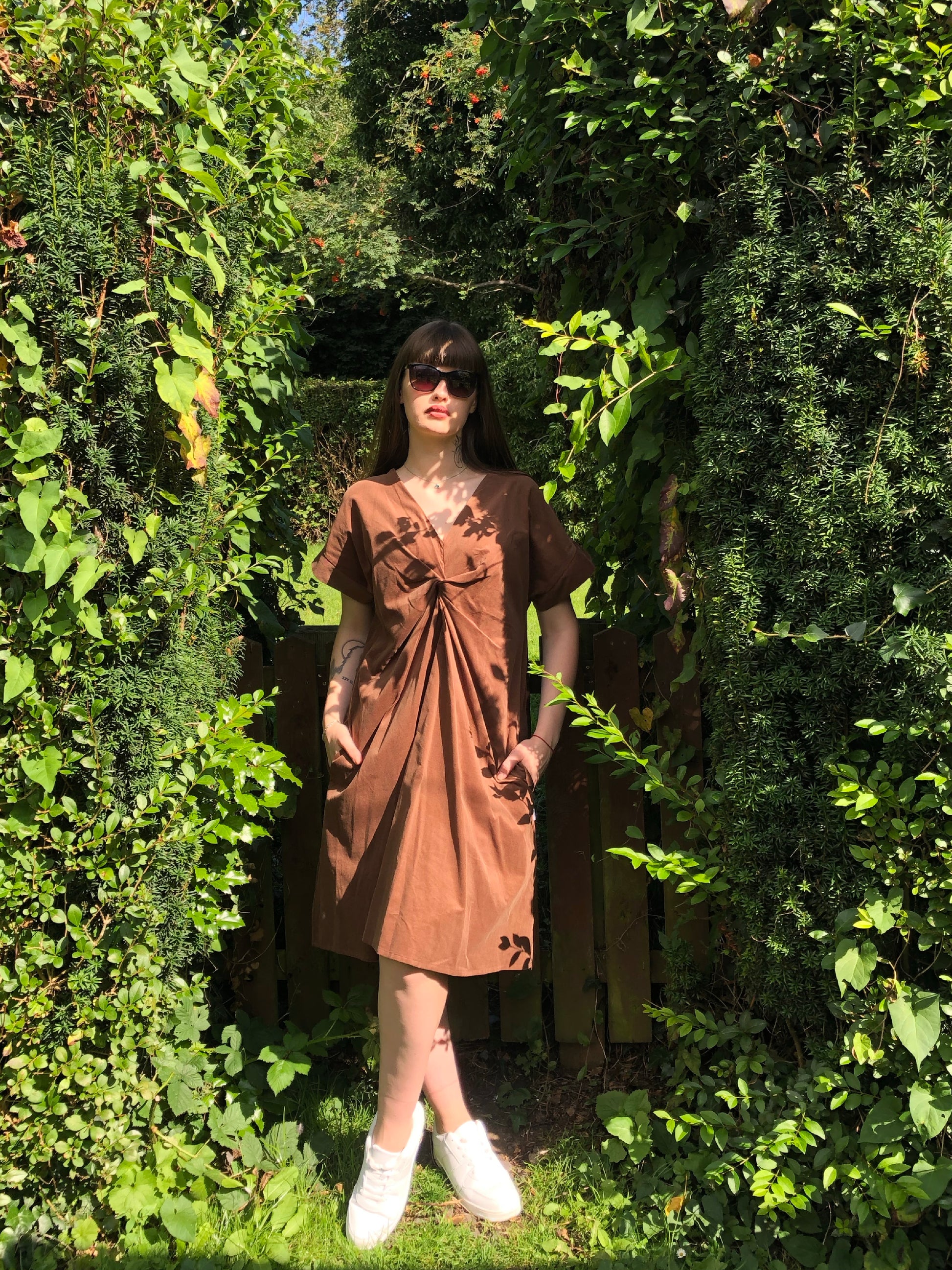 Pictured is the Riverbank Dress With a Knot in Brown Corduroy. Our model is standing against a fence in the woodlands. It is a Midi length dress, worn with white trainers