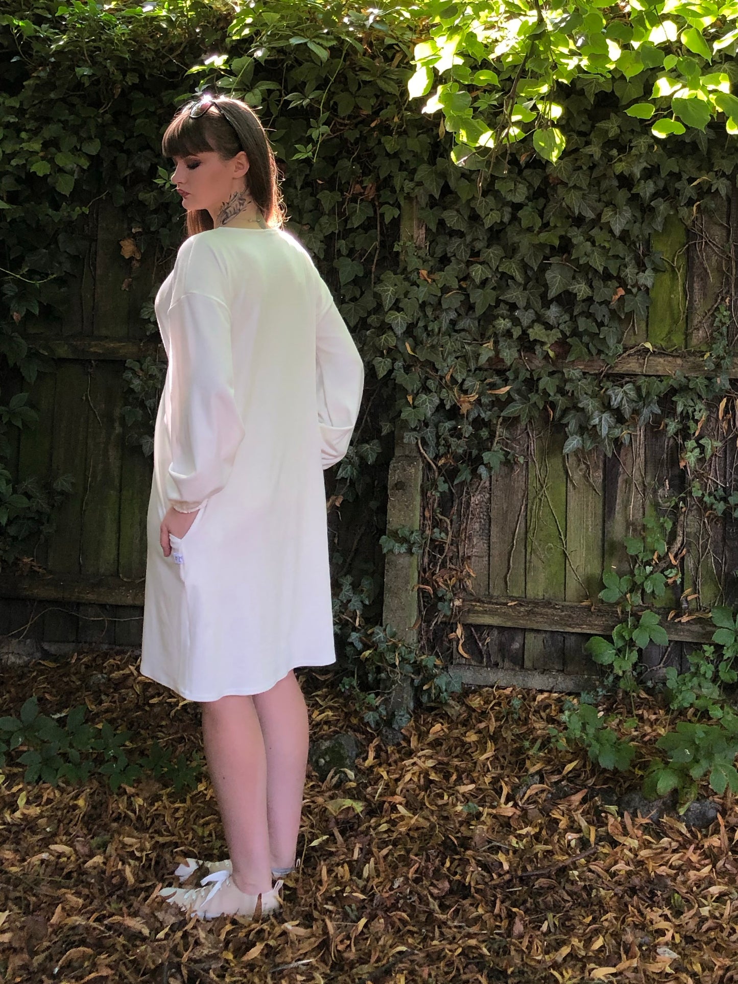 Our model has long dark hair and has her back to the camera. She is wearing a winter white long sleeved ribbed shift dress with pockets against a fenced background with foliage. The sleeves are elasticated at the cuff.