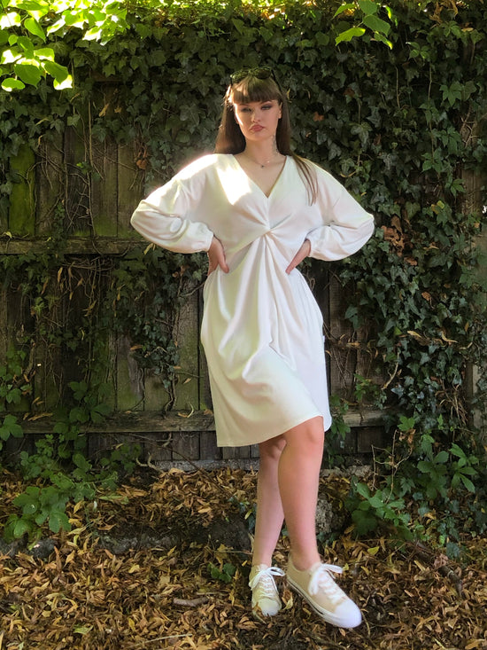 Our model has long dark hair and is facing the camera. She is wearing a winter white long sleeved ribbed shift dress with pockets against a fenced background with foliage. It has a plunge v neck and centre front knot. The sleeves are elasticated at the cuff.