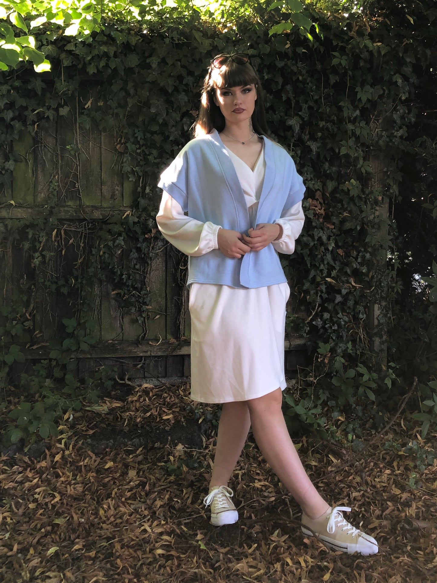 Here our model with brown hair is wearing the Signature Twilight Shrug in Baby Blue on top of the Meander Dress in Winter White. Paired with Beige trainers