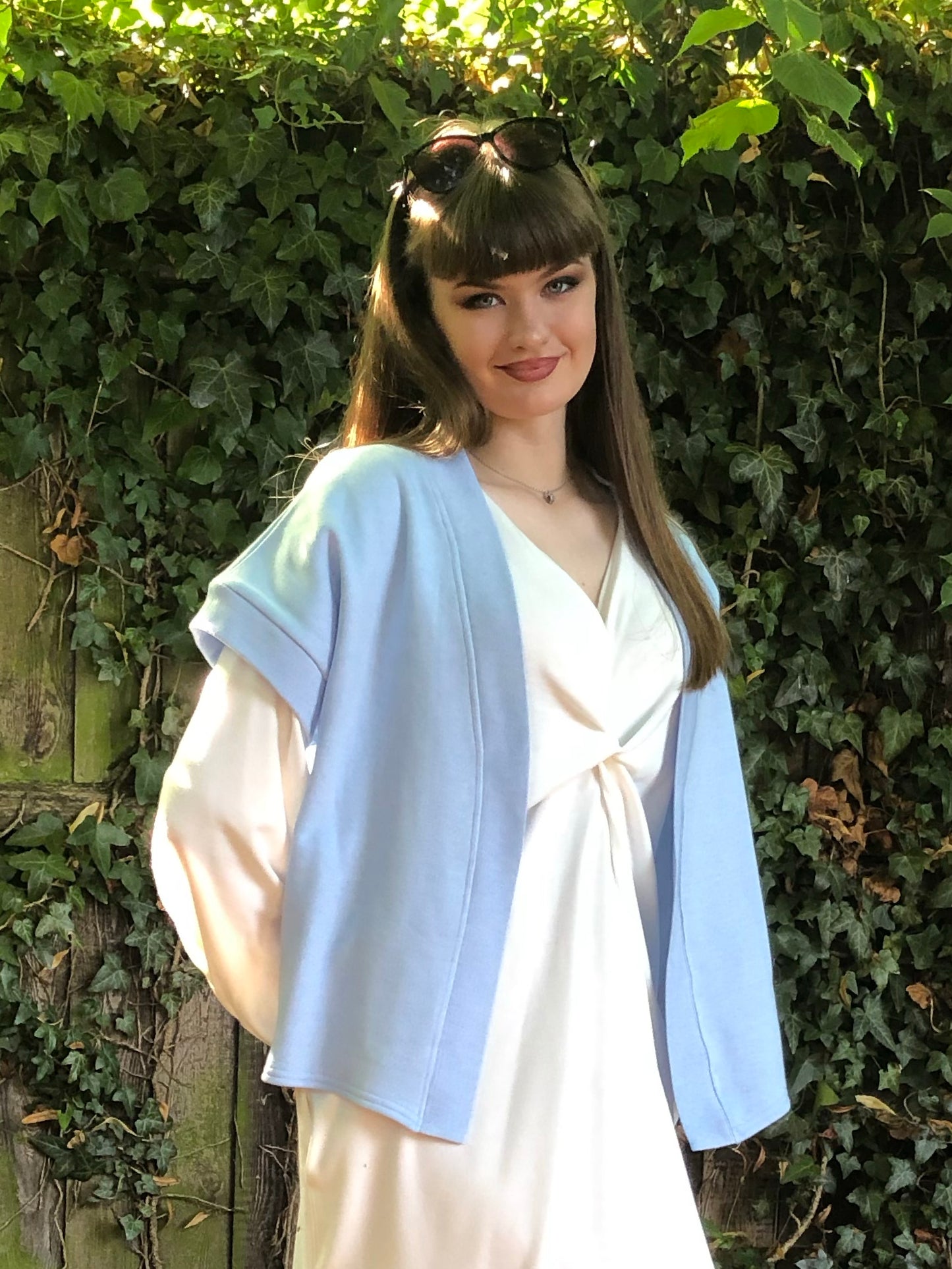 Here is a close up of our model with brown hair is wearing the Signature Twilight Shrug in Baby Blue on top of the Meander Dress in Winter White. Paired with Beige trainers