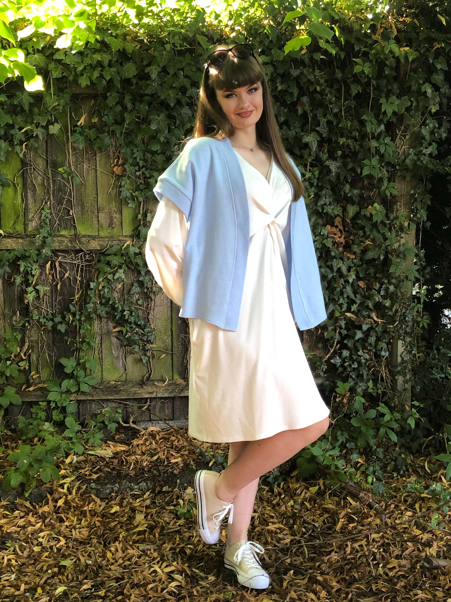 Our model has long dark hair and is facing the camera. She is wearing a winter white long sleeved ribbed shift dress with pockets against a fenced background with foliage. Over the top of the dress she is wearing our baby blue Signature Shrug. The dress has a plunge v neck and centre front knot. The sleeves are elasticated at the cuff.