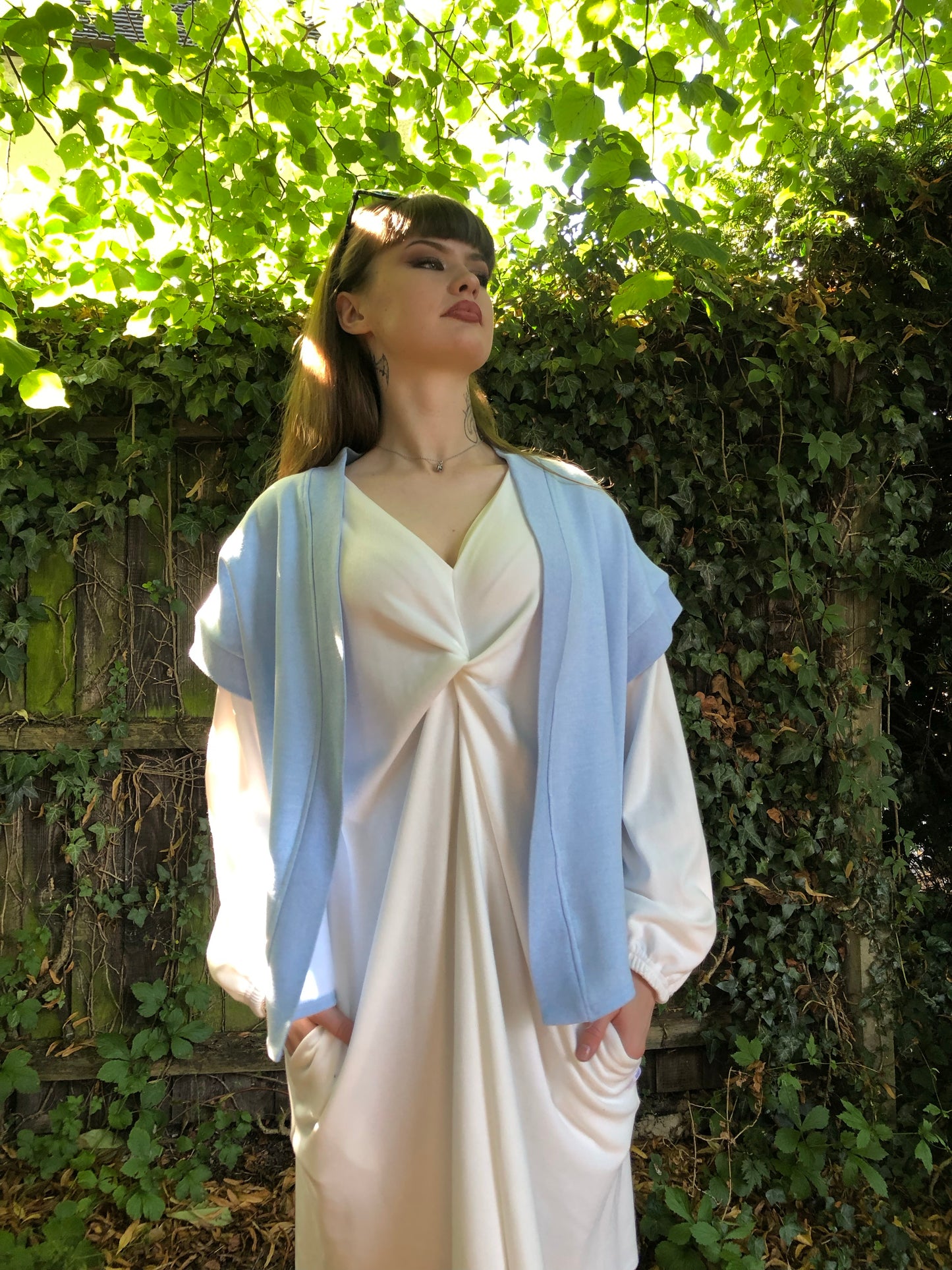 Here our model with brown hair is wearing the Signature Twilight Shrug in Baby Blue on top of the Meander Dress in Winter White. Paired with Beige trainers