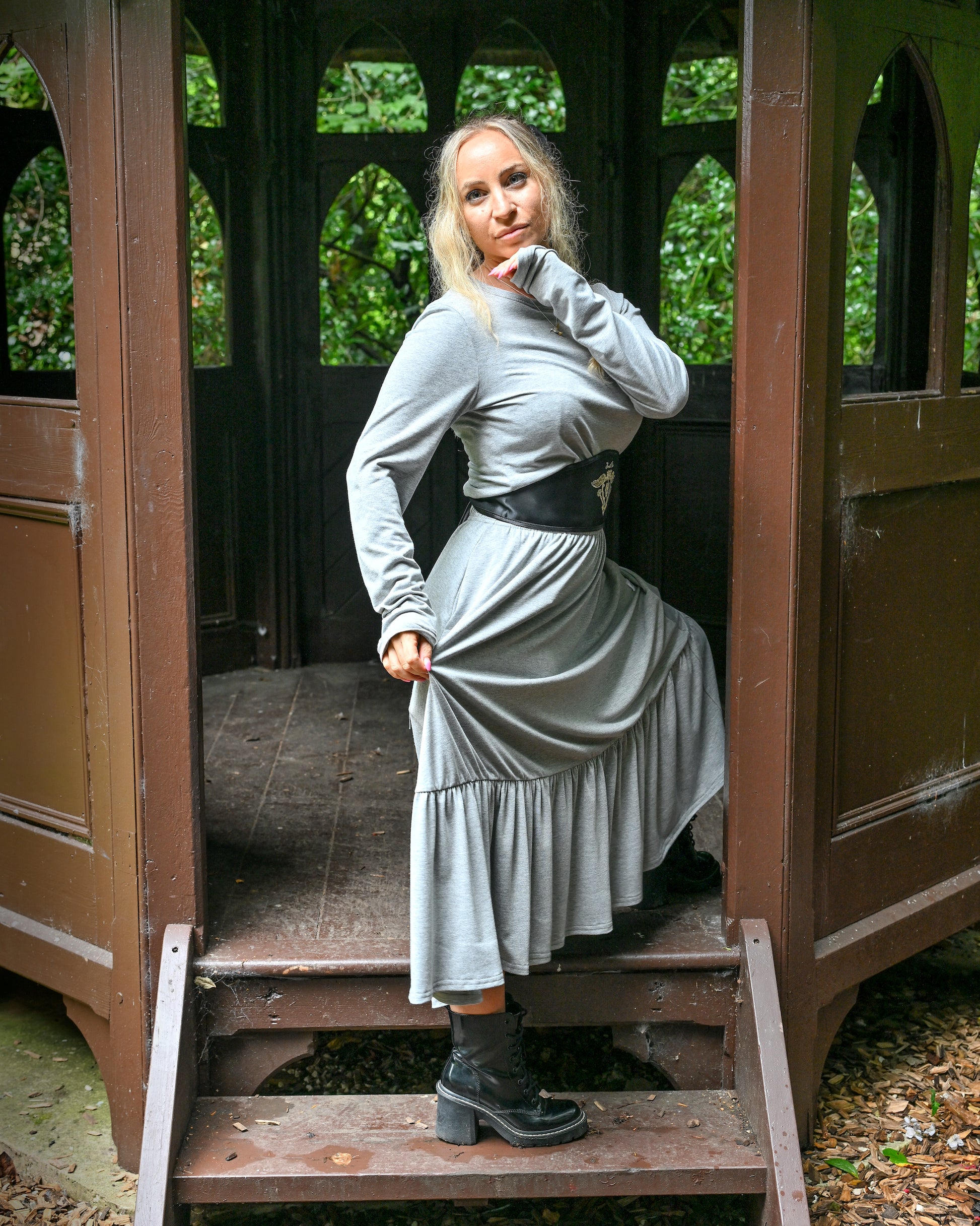 This is a young woman wearing a grey tiered skirt with a matching grey long sleeve. She is wearing a black leather under-bust style belt and leather heeled boots. She is silhouetted against a wooden structure, stood on the steps. She has one hand under her chin and another holding and extending the skirt.