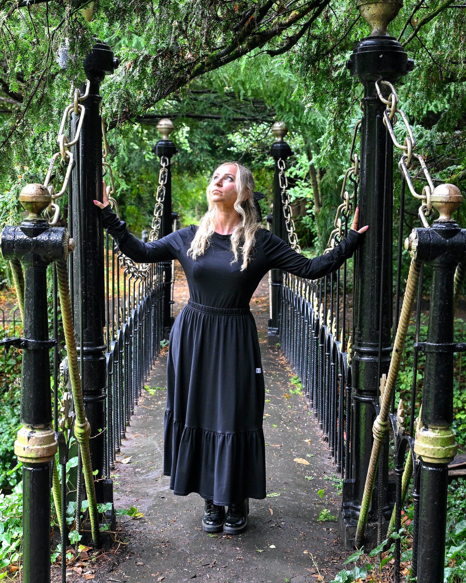 The young woman in the picture has long blonde hair and she is standing on a bridge in a park with lots of trees and foliage. She is holding onto the poles at the entrance to the bridge and is glancing upwards to her right and is facing the camera. She is wearing a boho gathered midi skirt with pockets in black. She is also wearing our beatnik top with crew neck in black. The top has ruched sleeves below the elbows. She is wearing black boots.