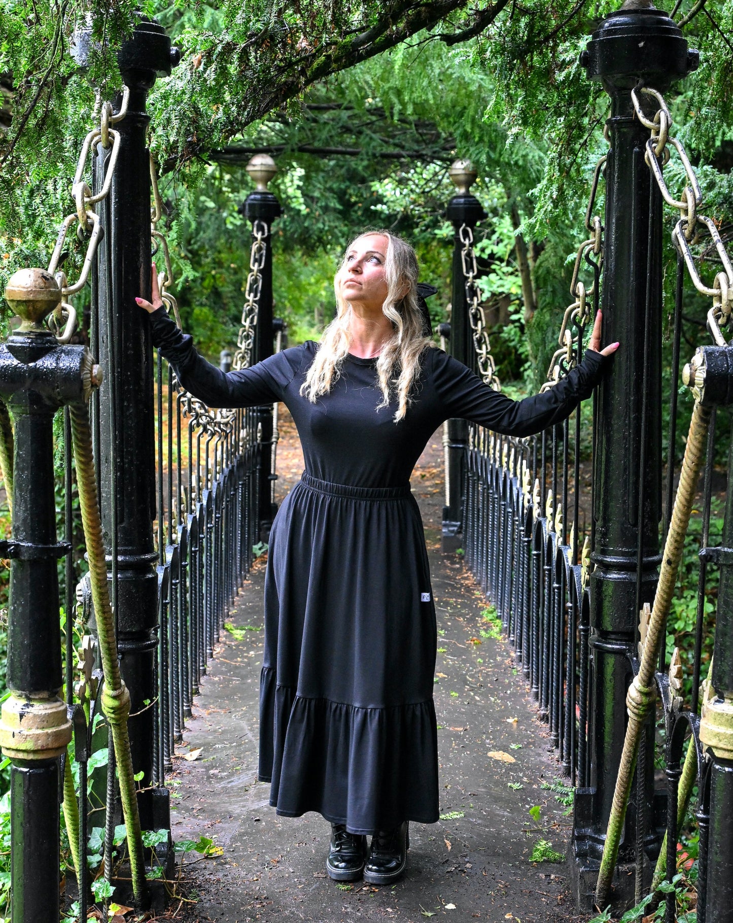 The young woman in the picture has long blonde hair and she is standing on a bridge in a park with lots of trees and foliage. She is holding onto the poles at the entrance to the bridge and is glancing upwards to her right and is facing the camera. She is wearing a beatnik top with crew neck in black. The top has ruched sleeves and is being worn with our boho black skirt and black boots.