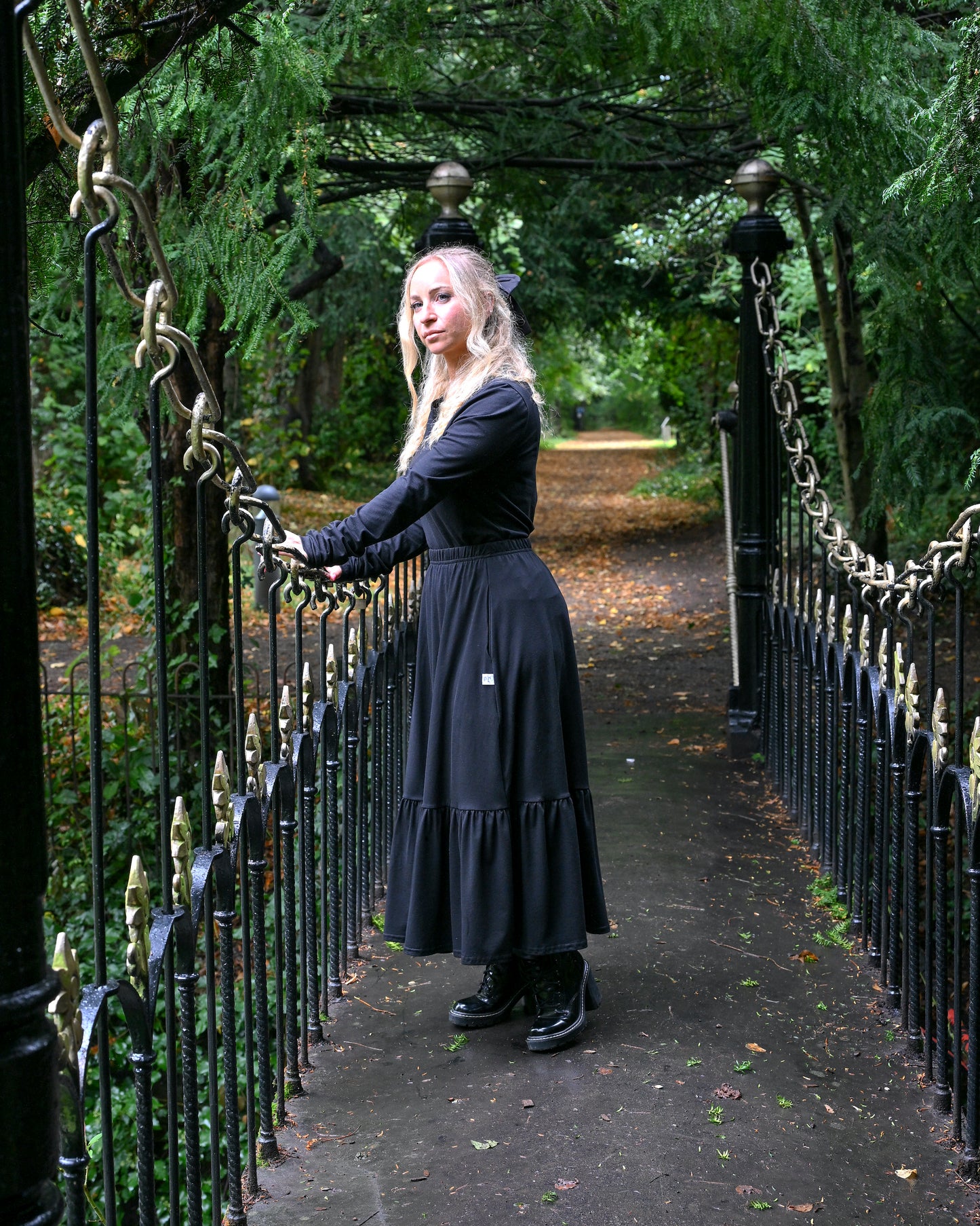 The young woman in the picture has long blonde hair and she is standing in the middle of a small bridge in a park with lots of trees and foliage. She is holding onto the railings of the bridge with both hands and is facing the camera. The bridge is very ornate and is black and gold in colour. She is wearing a boho gathered midi skirt with pockets in black. She is also wearing our beatnik top with crew neck in black. The top has ruched sleeves below the elbows. She is wearing black boots.