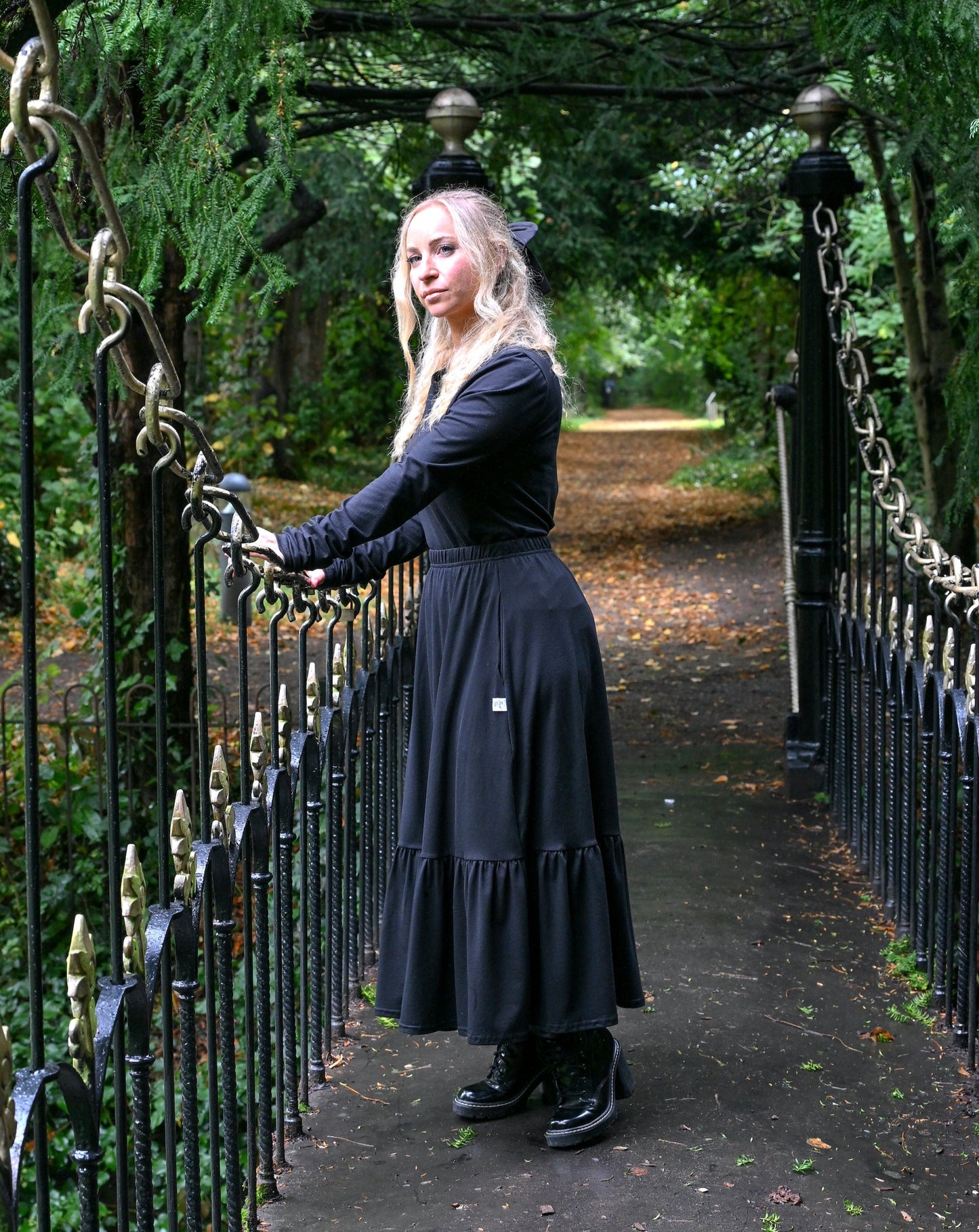 The young woman in the picture has long blonde hair and she is standing in the middle of a small bridge in a park with lots of trees and foliage. She is holding onto the railings of the bridge with both hands and is facing the camera. The bridge is very ornate and is black and gold in colour. She is wearing a beatnik top with crew neck in black. The top has ruched sleeves and is being worn with our boho black skirt and black boots.