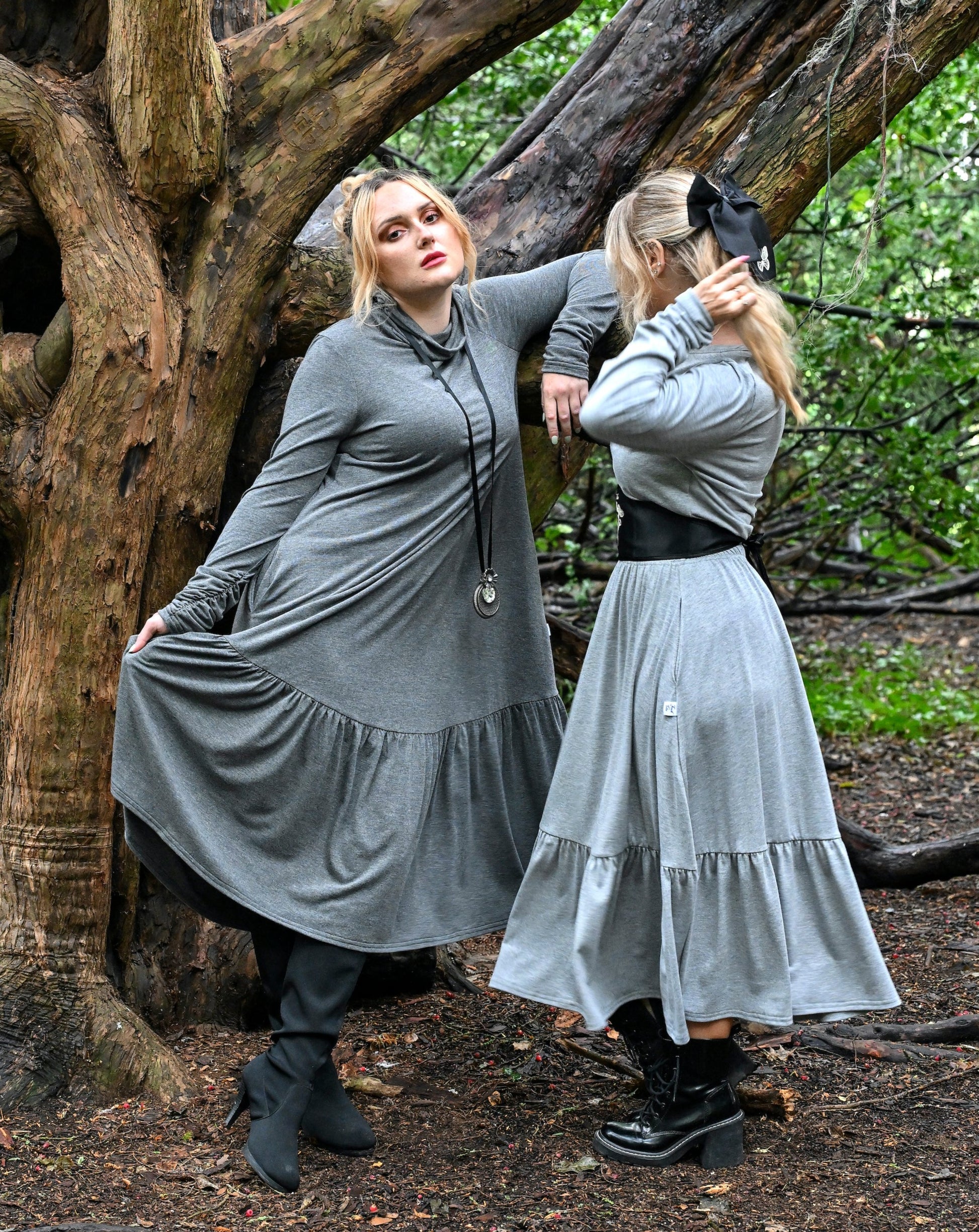 Two young ladies in a woodland area, one wearing a midi length grey dress with long sleeves and a loose neck, it has a gathered panel at the base of the skirt and ruched sleeves, she is wearing black heeled boots and a long necklace with a pendant. The other model is wearing a grey tiered skirt with a matching long sleeve t-shirt, she has a black leather under bust belt and leather heeled boots with a black bow in her hair.