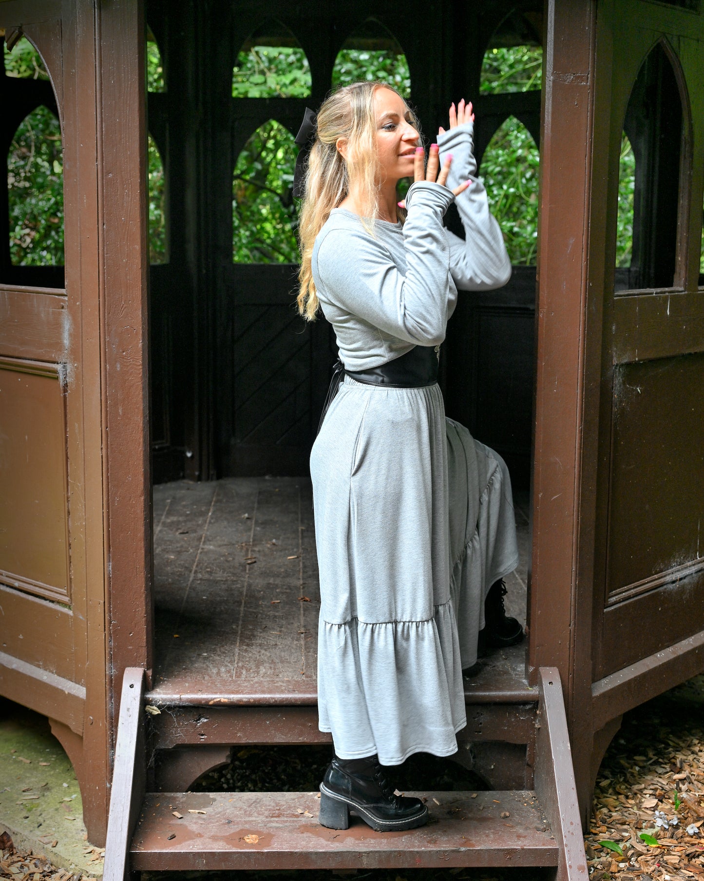 This is a young woman wearing a grey tiered skirt with a matching grey long sleeve. She is wearing a black leather under-bust style belt and leather heeled boots. She is silhouetted against a wooden structure, stood on the steps. She is stood to the side with her hands held out in front of her face.