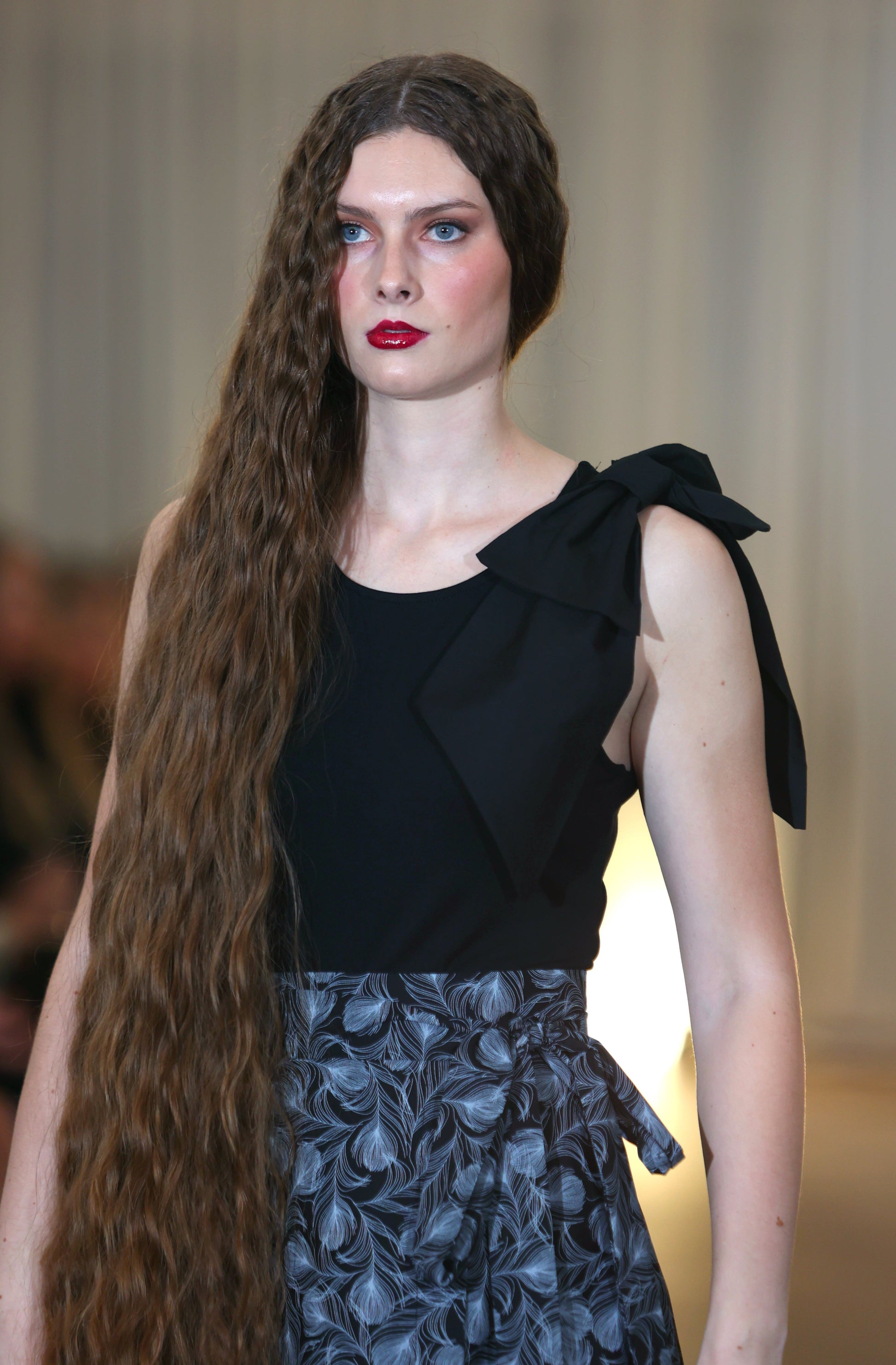 The young woman in the picture has very long dark hair and she is walking towards the camera. She has blue eyes and is wearing red lipstick. She is wearing a brook top with bow in black skirt and a boho gathered midi skirt in navy print. Both hands are by her side. 