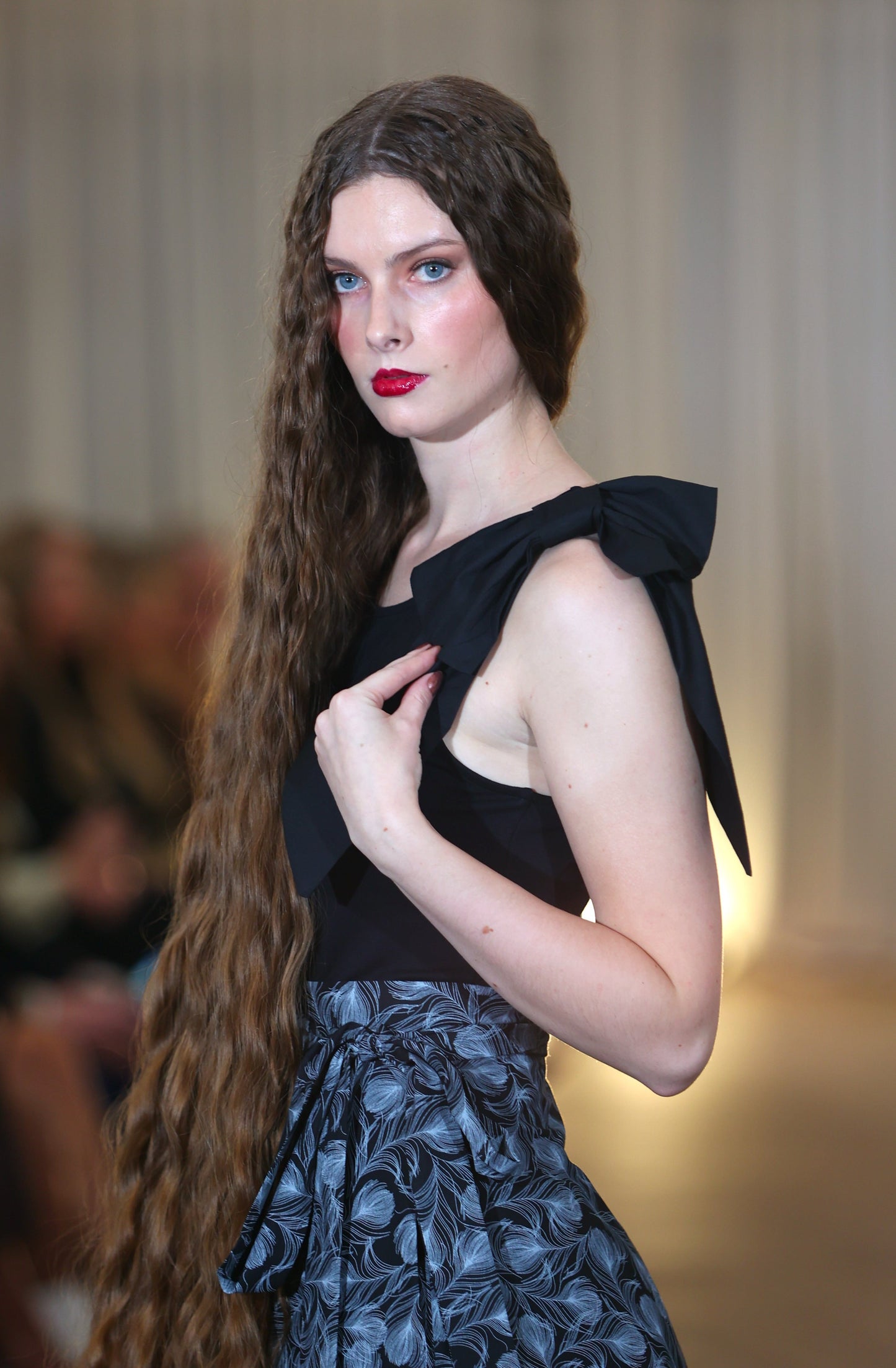 The young woman in the picture has very long dark hair and she is facing the camera. She has blue eyes and is wearing red lipstick. She is wearing a brook top with bow in black skirt and a boho gathered midi skirt in navy print. Her left hand is holding the bow.