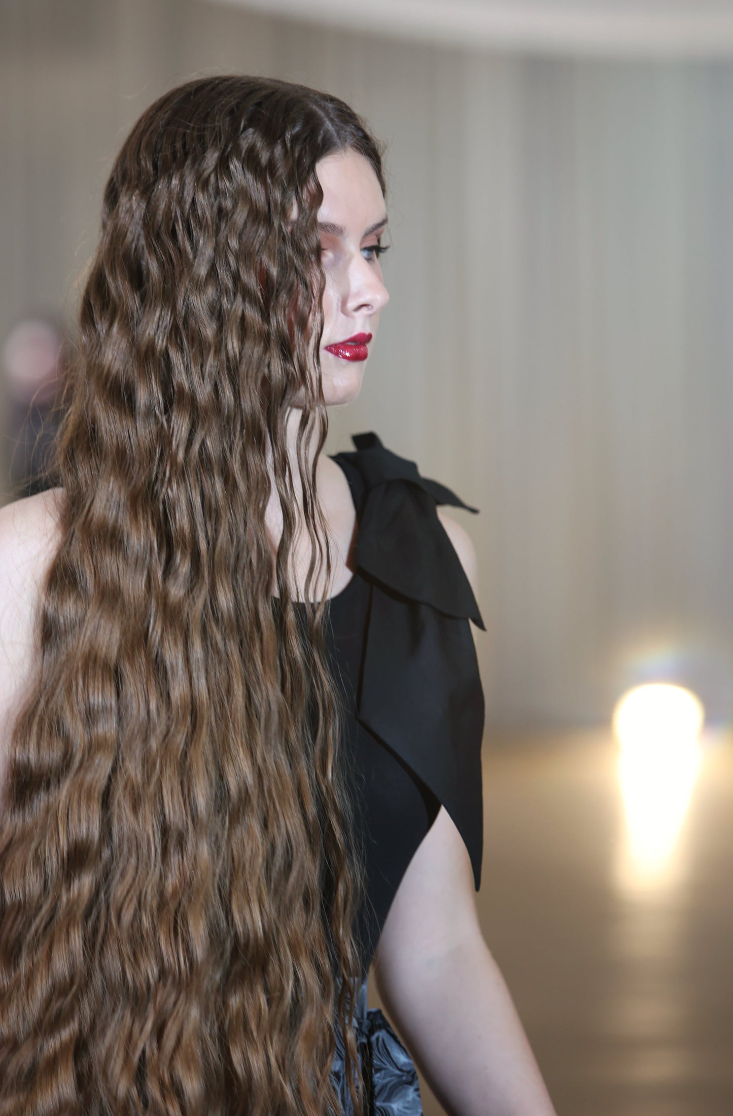 The young woman in the picture has very long dark hair and she has the right side of her face to the camera. She is wearing red lipstick. She is wearing a brook top with bow in black. Her left hand is by her side.