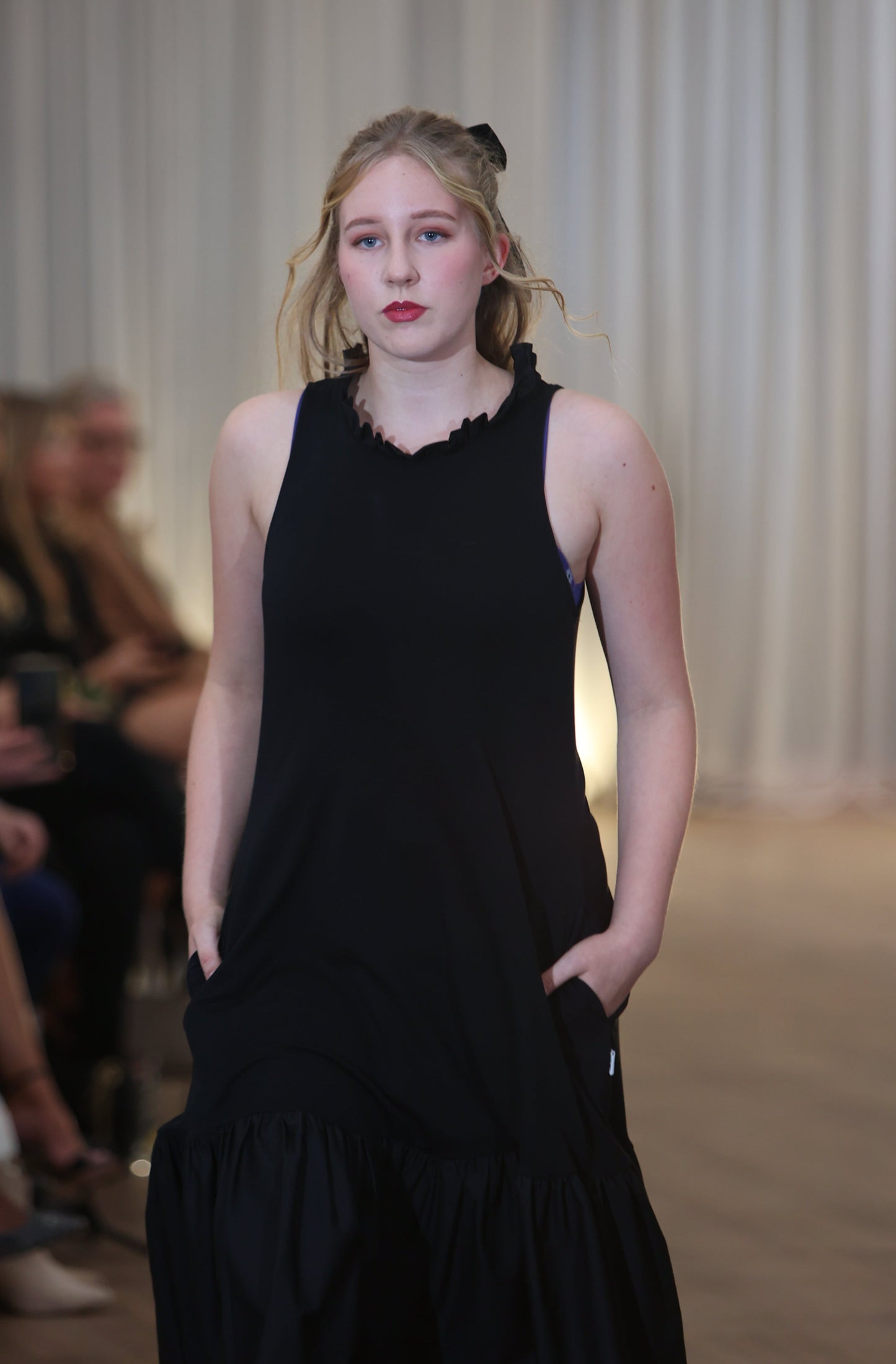 There is a young woman walking towards the camera in a crowded room with long white curtains in the background. She has long blonde hair tied in a bow. She is wearing the iris midi dress with pockets in black. The dress has a high neck with a row of ruffles. Both hands are in her pockets.