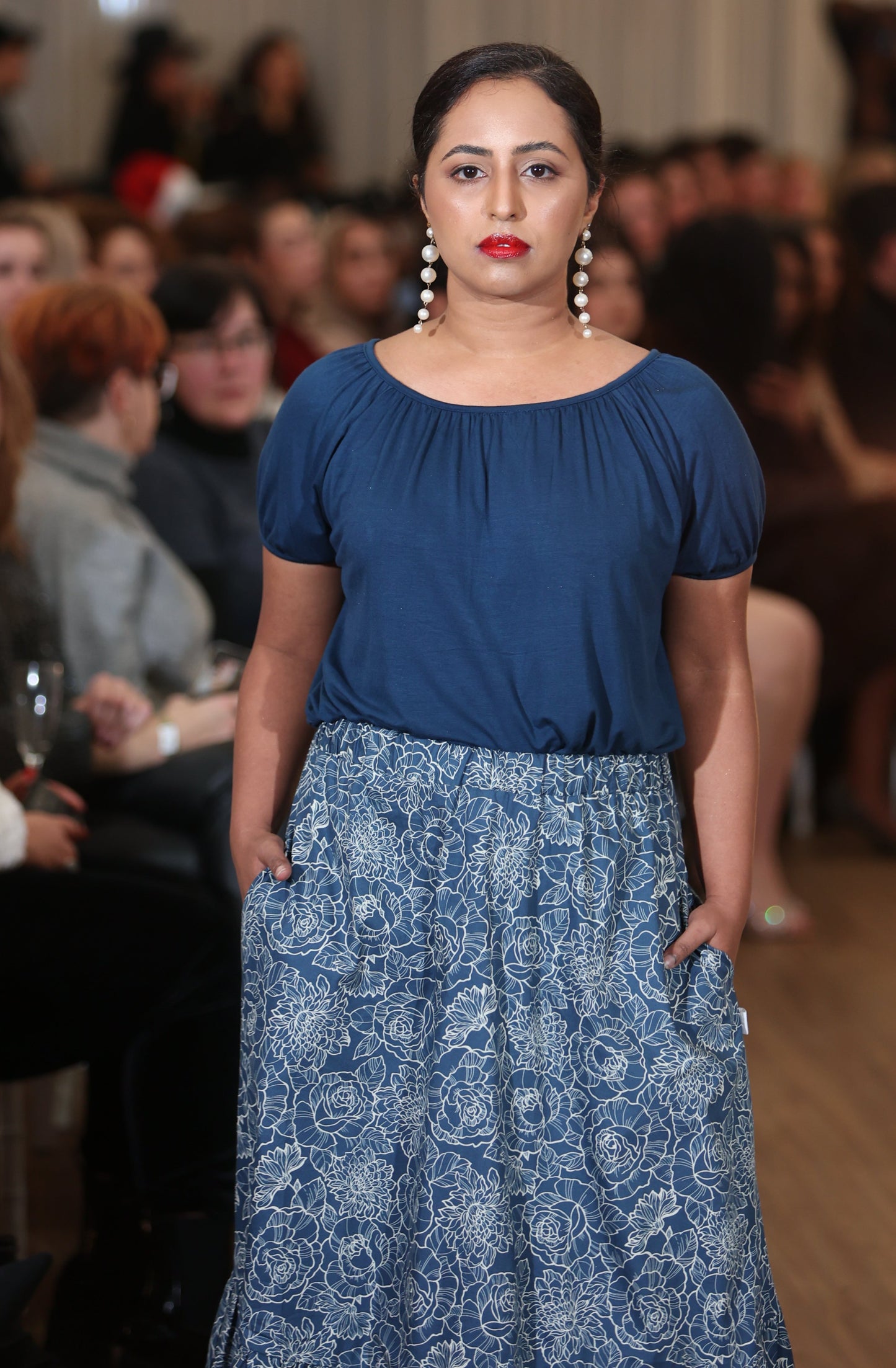 The young woman in the picture has dark hair and she is walking towards the camera in a crowded room. She has long silver earrings and red lipstick. She is wearing a boho gathered midi skirt with pockets in navy print. She is also wearing our posey top in french navy. Both hands are in her pockets.  