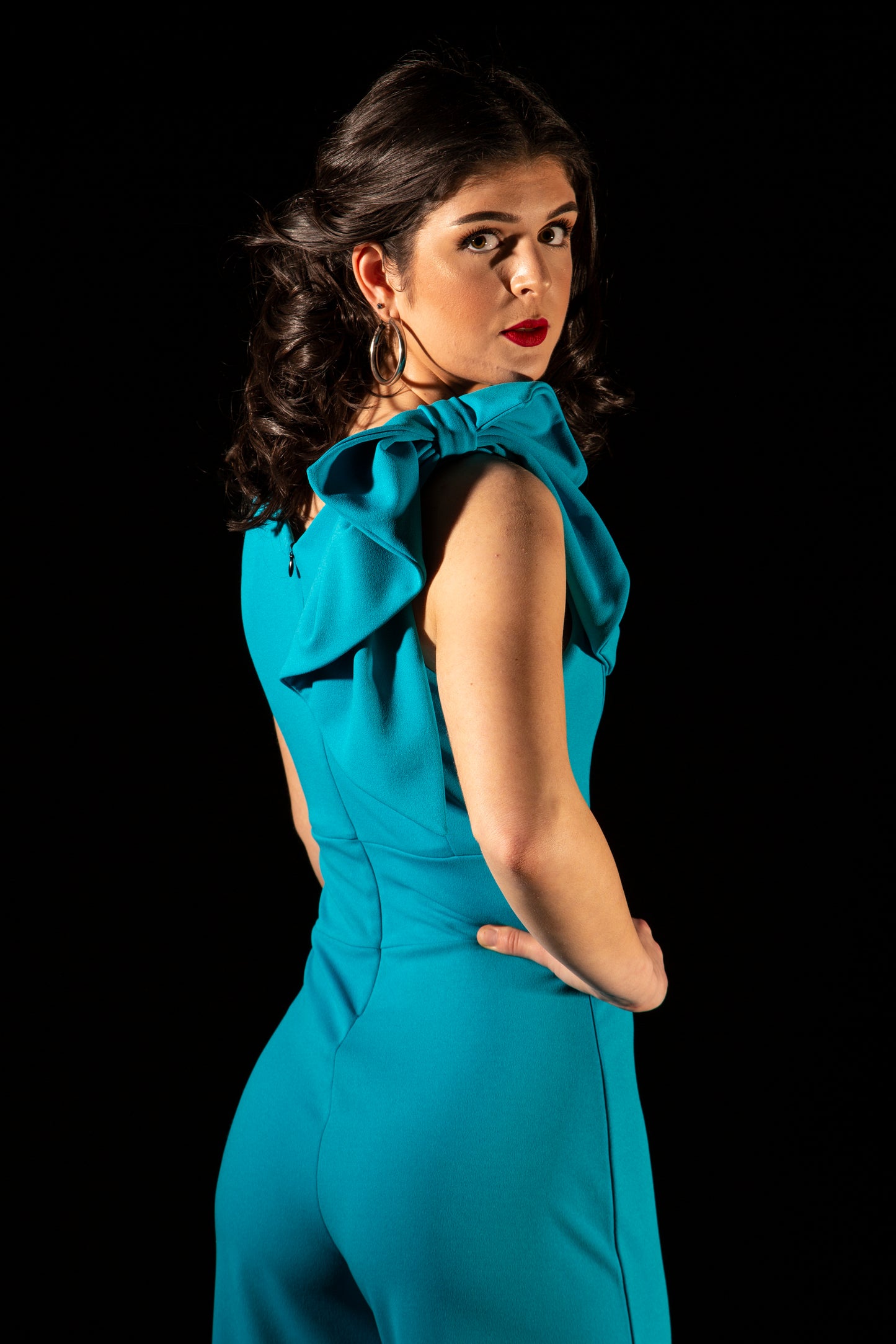 This is a young lady wearing a Turquoise colour V neck jumpsuit with wide legs and a bow on the shoulder. She is standing with her hand on her hip, looking over her shoulder, silhouetted against a black wall.