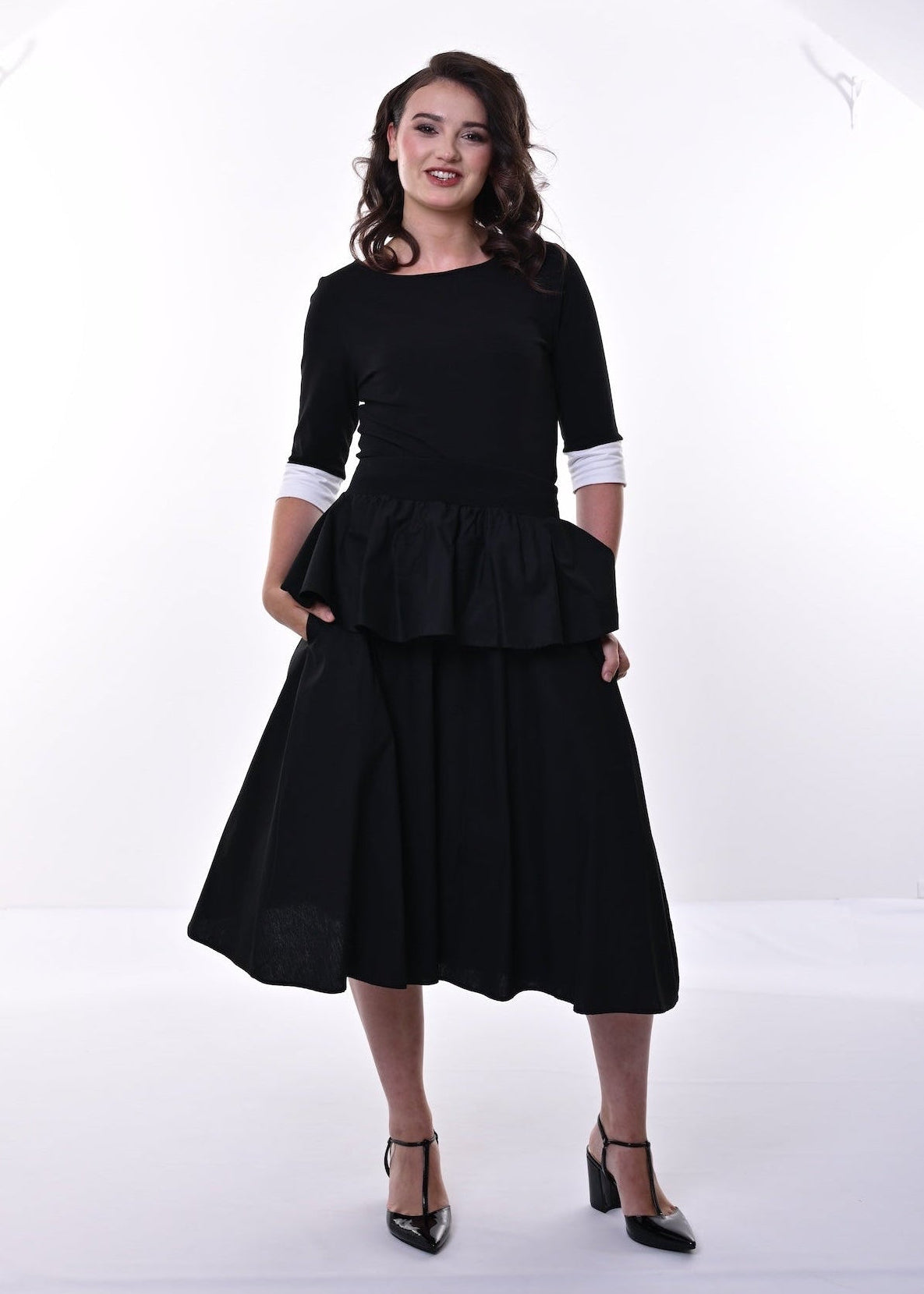 A woman modelling a black top and skirt with a peplum waist, 3/4 sleeves, and contrasting cream cuffs, paired with black heeled sandals, set against a plain white studio background.