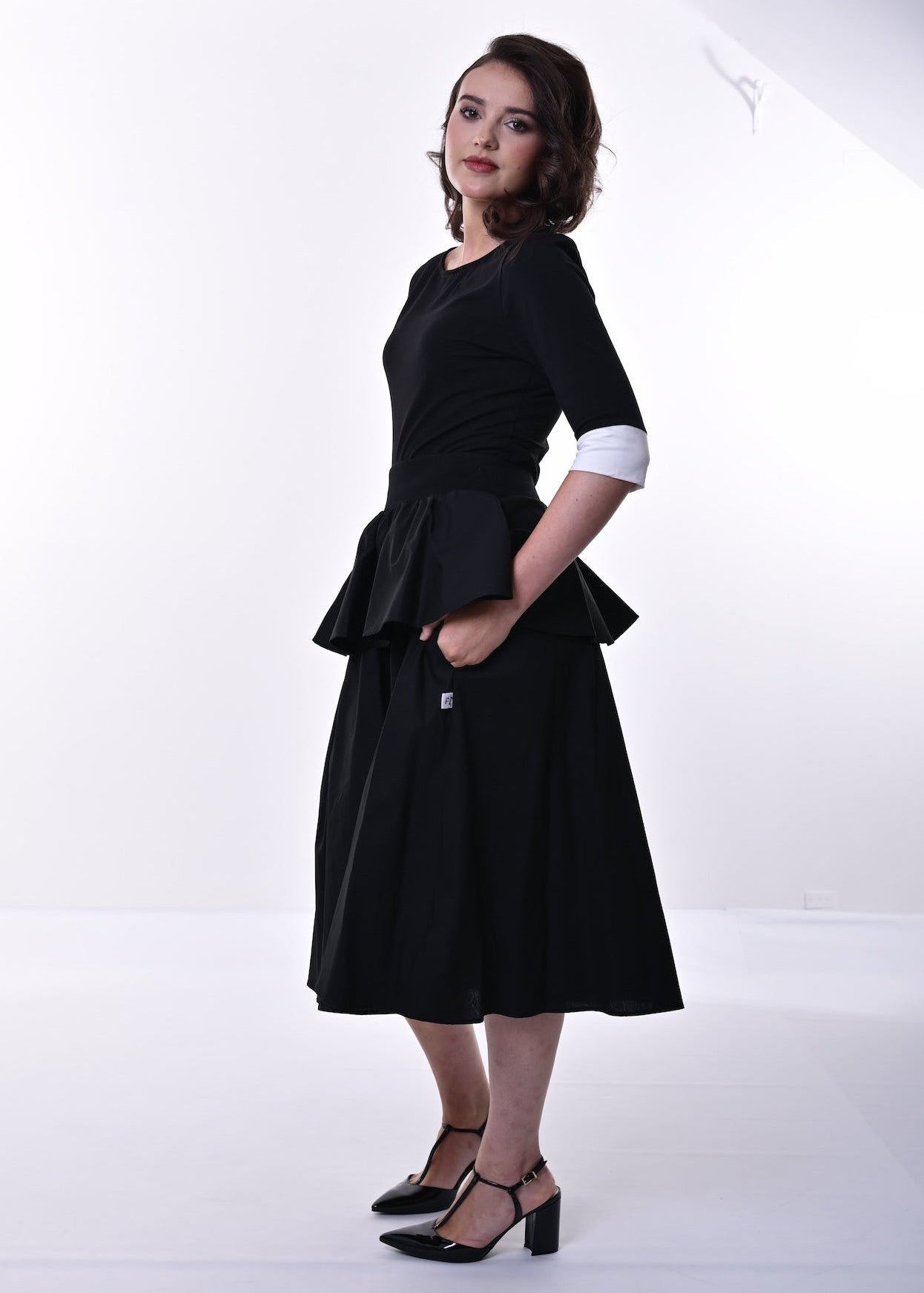 A woman modelling a black top and skirt with a peplum waist, 3/4 sleeves, and contrasting cream cuffs, paired with black heeled sandals, set against a plain white studio background.