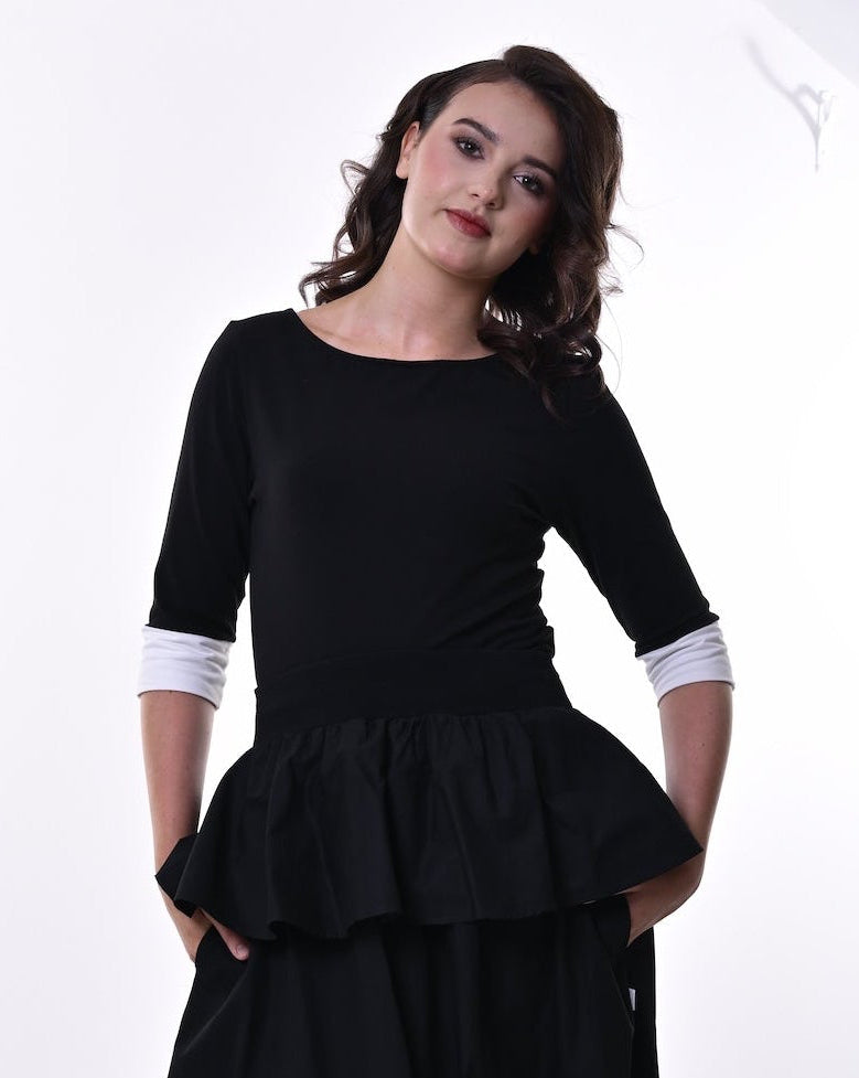 A woman modelling a black top and skirt with a peplum waist, 3/4 sleeves, and contrasting cream cuffs, paired with black heeled sandals, set against a plain white studio background.