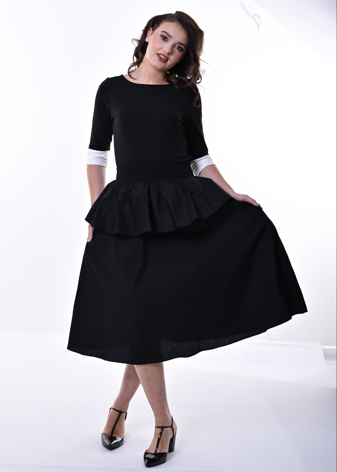 A woman modelling a black top and skirt with a peplum waist, 3/4 sleeves, and contrasting cream cuffs, paired with black heeled sandals, set against a plain white studio background.