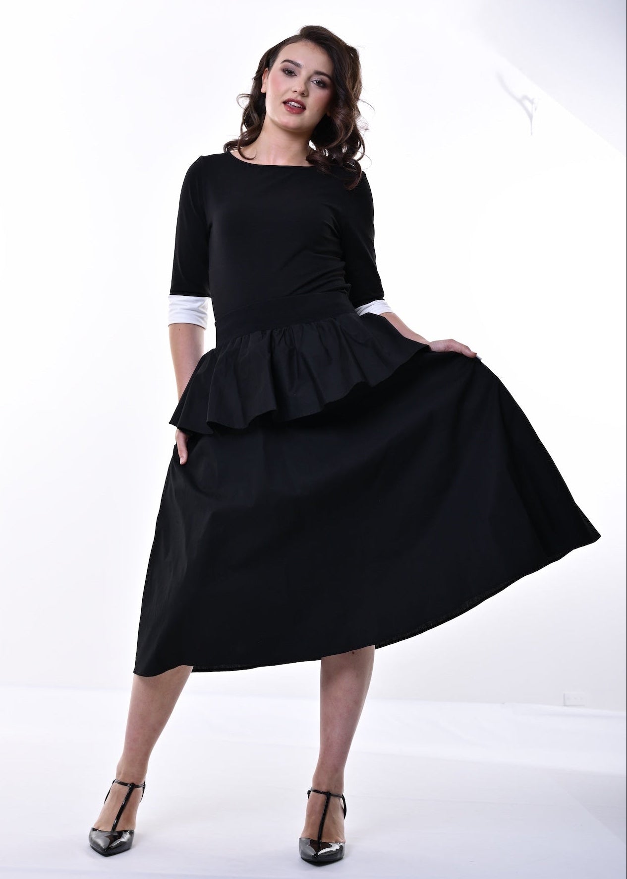A woman modelling a black top and skirt with a peplum waist, 3/4 sleeves, and contrasting cream cuffs, paired with black heeled sandals, set against a plain white studio background.