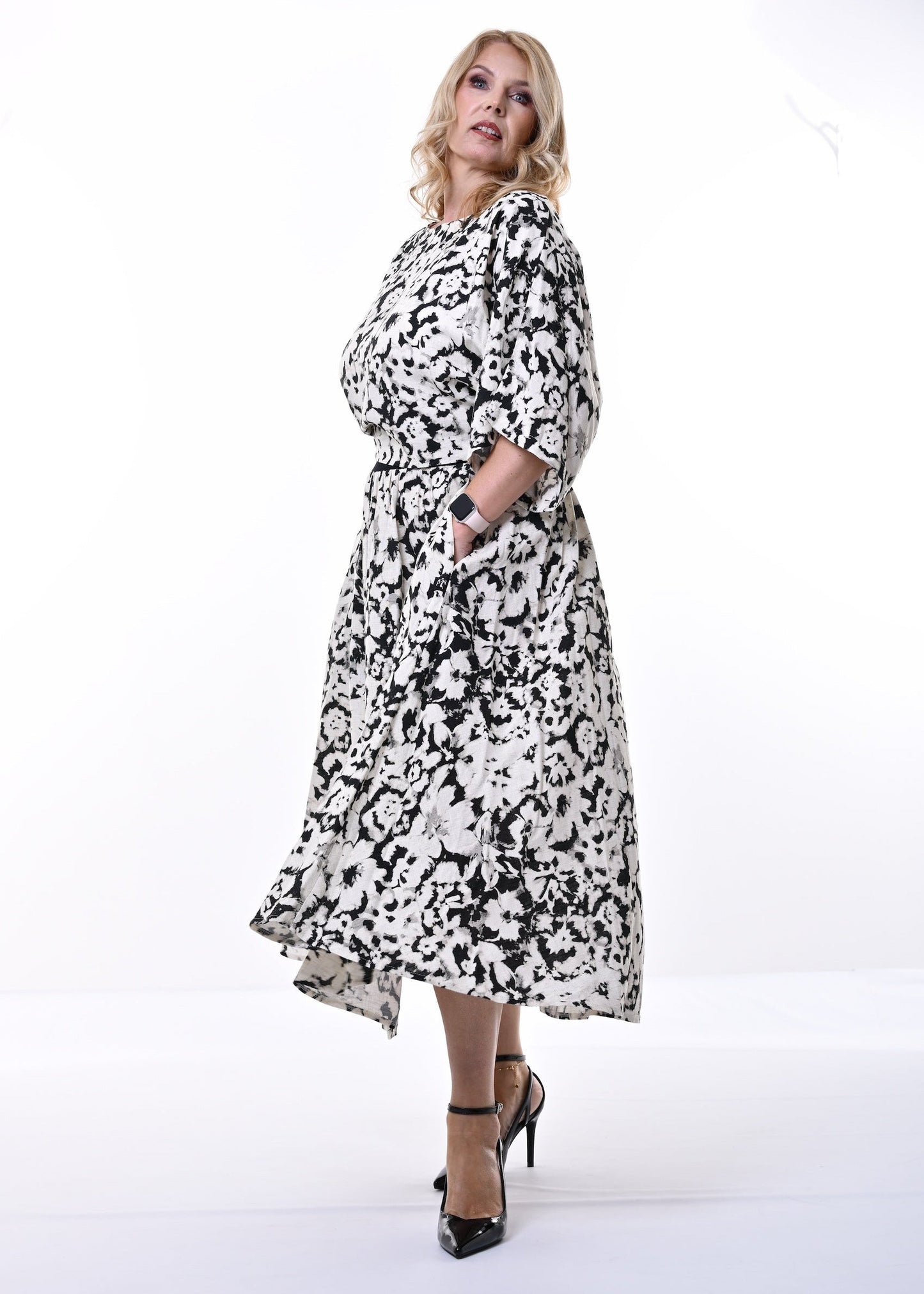 A woman modeling a black and white floral midi dress with three-quarter sleeves and a self-tie belt, standing against a plain white studio background.
