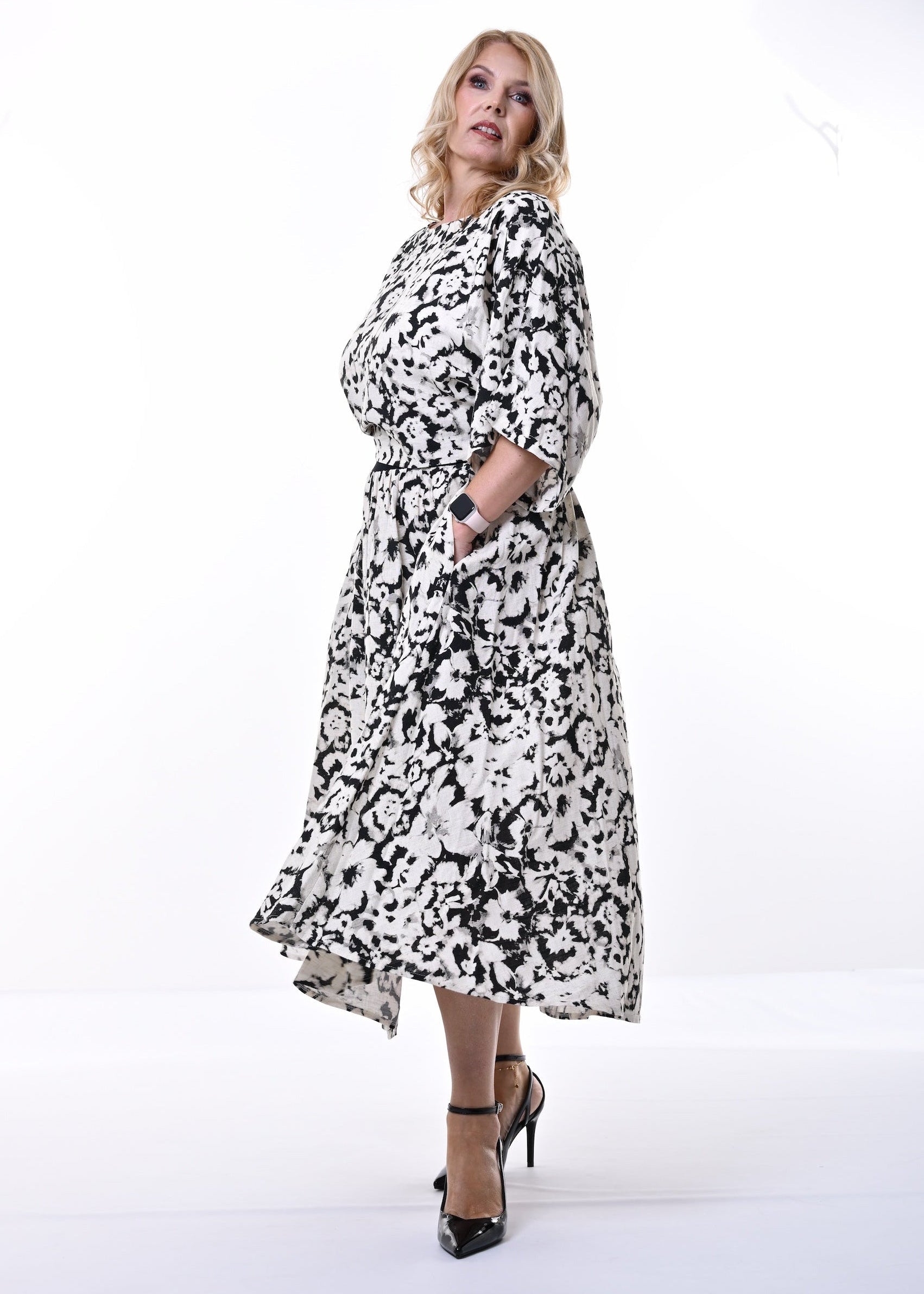 A woman modeling a black and white floral midi dress with three-quarter sleeves and a self-tie belt, standing against a plain white studio background.