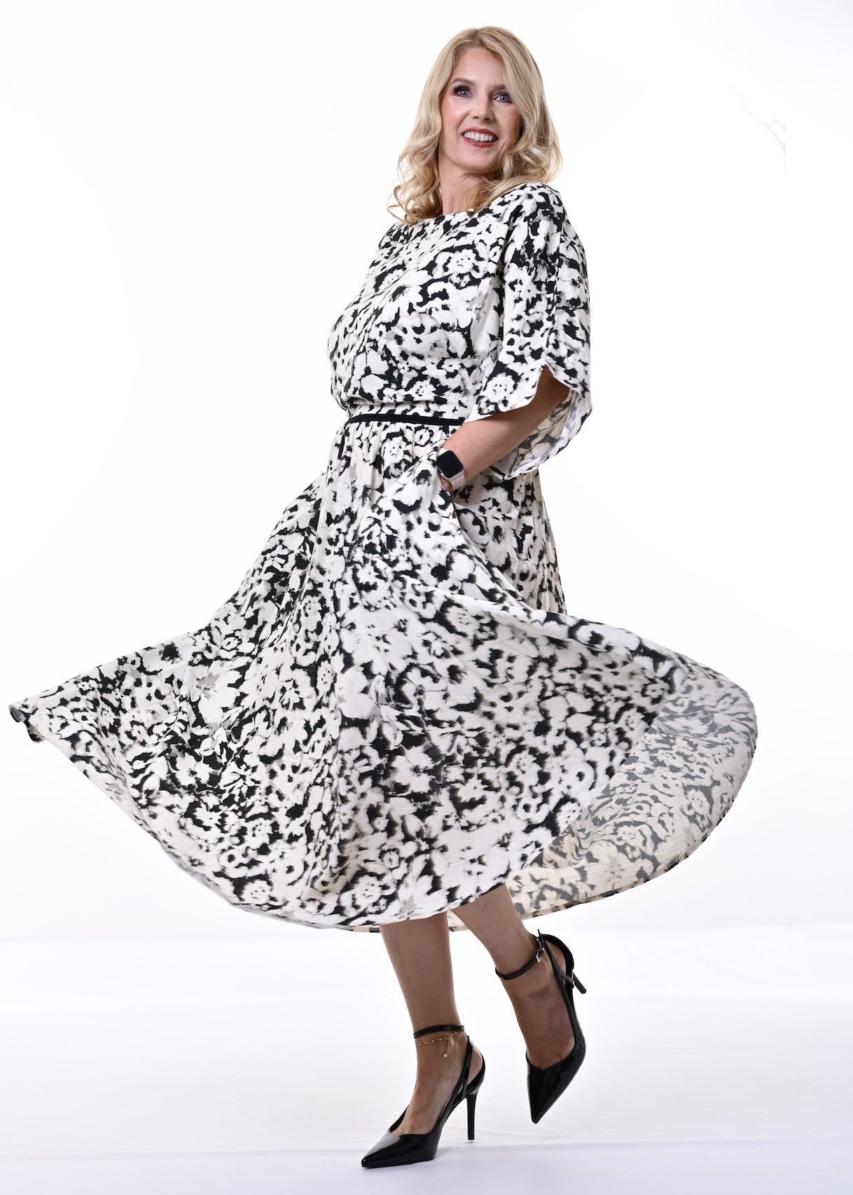 A woman modeling a black and white abstract patterned midi dress with three-quarter sleeves and a matching belt, captured in motion as she twirls on a bright white studio background.