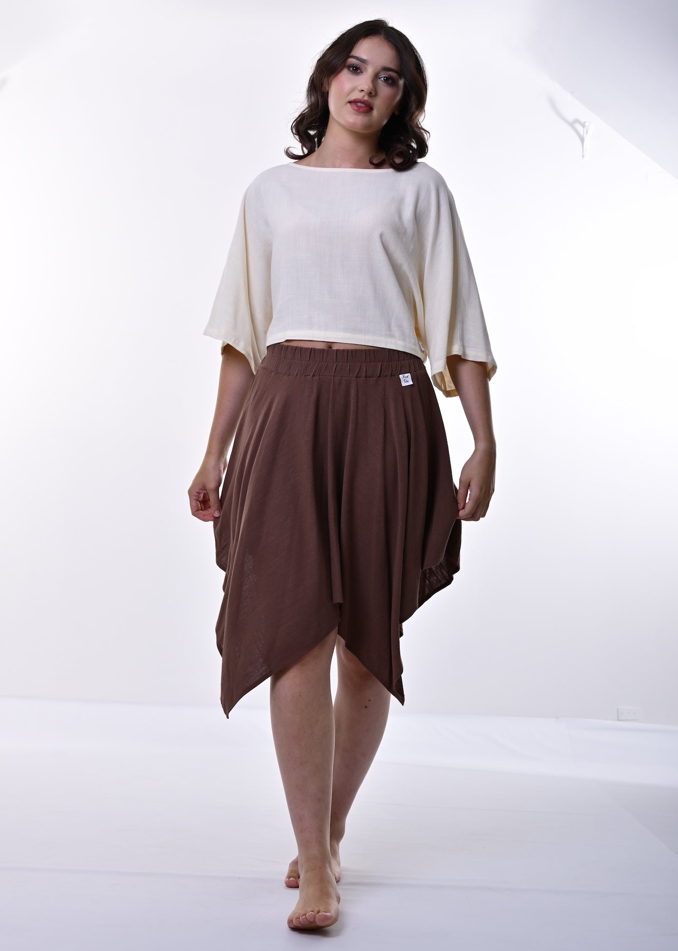 A woman modeling a cream-colored short-sleeve top and a brown draped midi skirt against a bright white studio background.