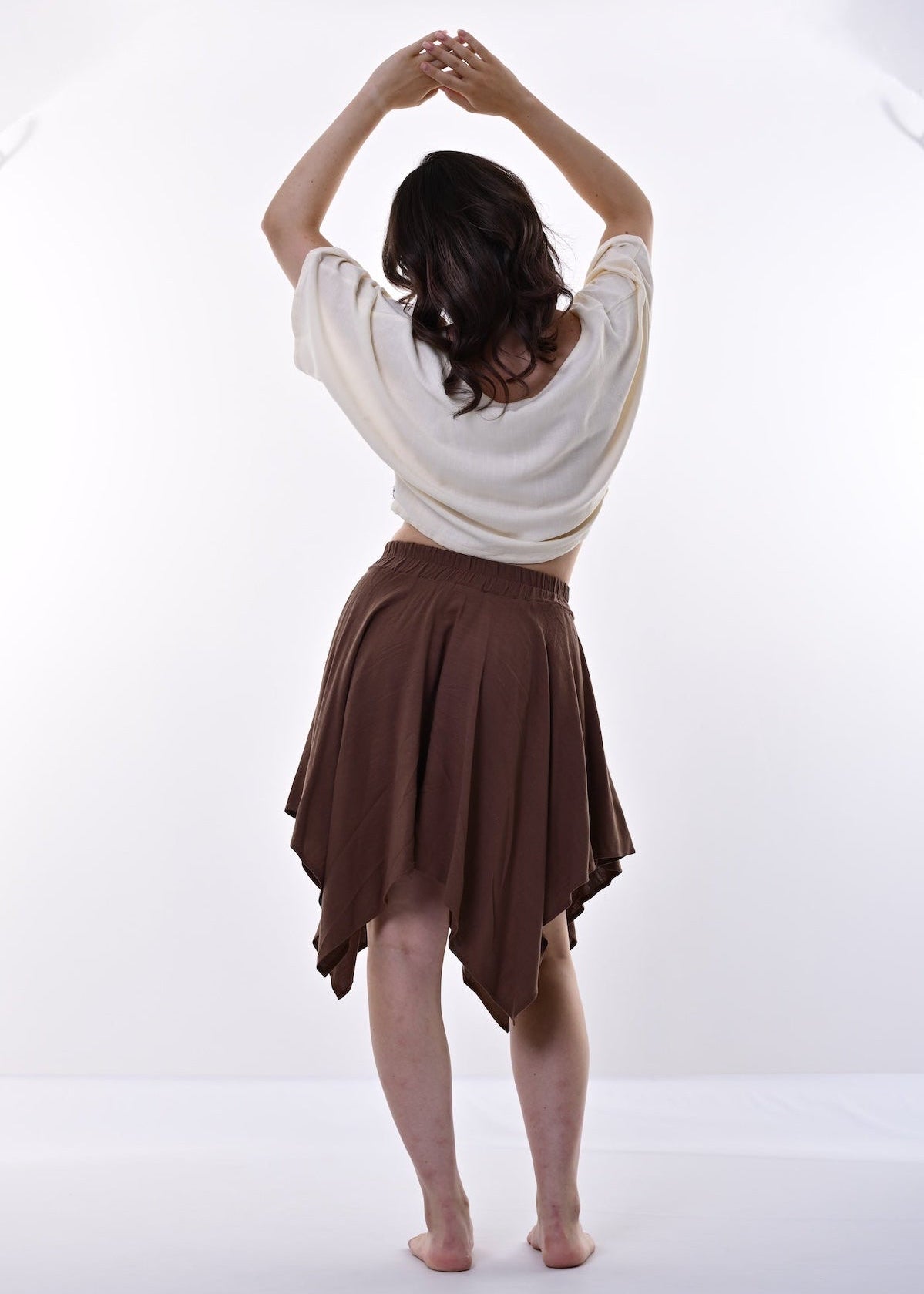 A woman seen from behind wearing a cream-colored short-sleeve crop top and a brown hanky hem skirt, standing barefoot against a bright white studio background.