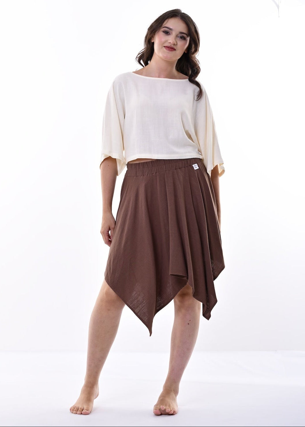A woman modeling a two-piece outfit consisting of a cream-colored short-sleeve top and a brown hanky hem skirt, standing against a bright white studio background.